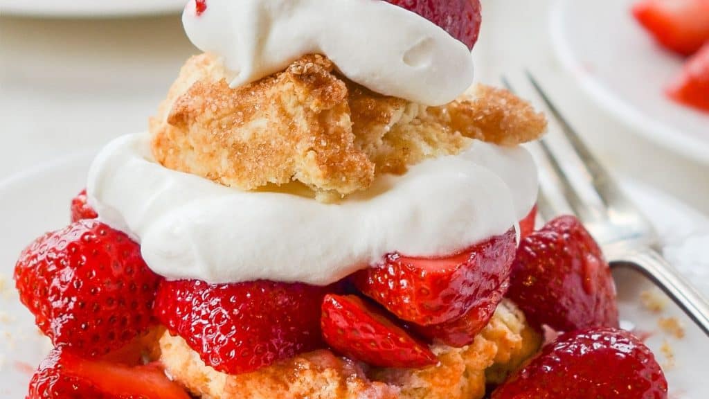 A golden biscuit topped with fresh, sliced strawberries and layers of fluffy whipped cream. The dessert is stacked high, with another biscuit layer and a final dollop of whipped cream crowning the treat.
