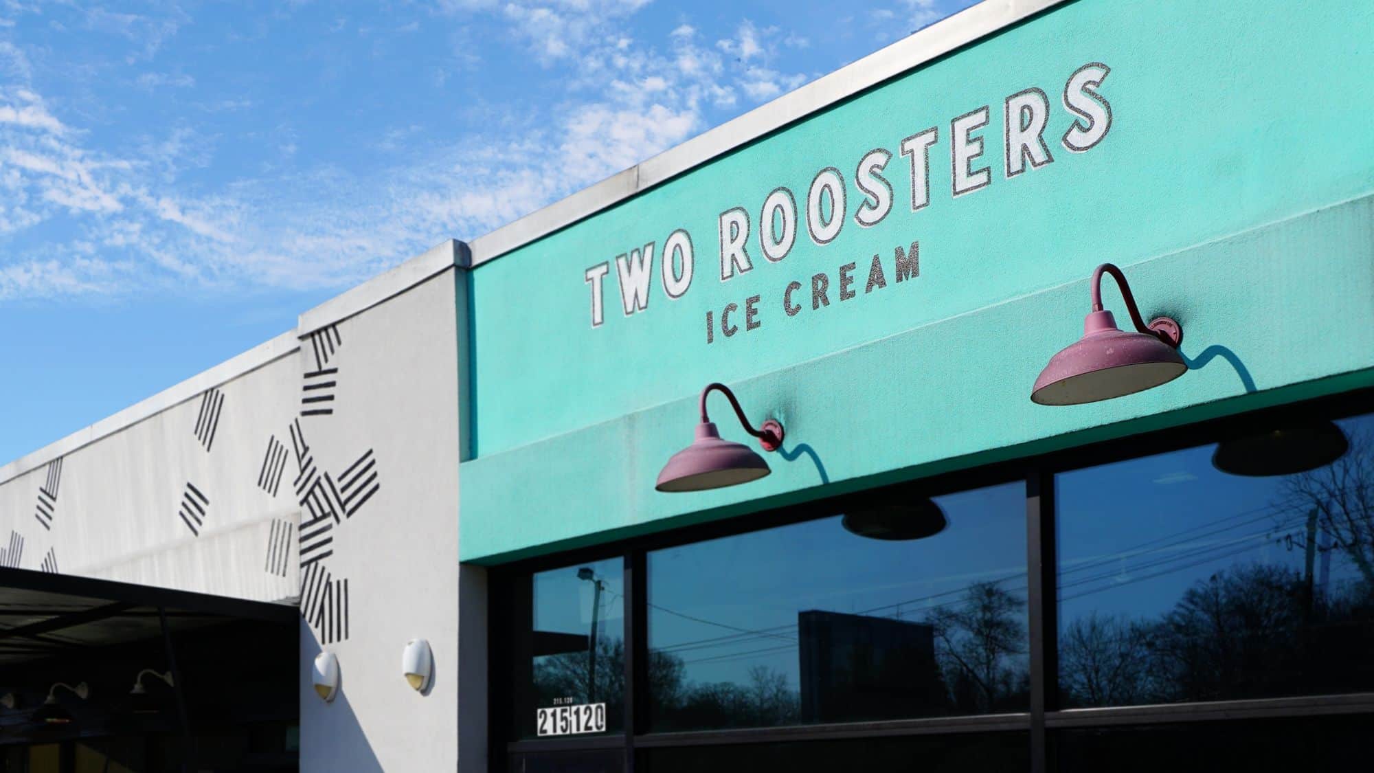 A storefront of Two Roosters Ice Cream with a turquoise and white exterior featuring bold signage and pink industrial-style lamps. The bright sky and reflections in the large windows add to the inviting atmosphere.