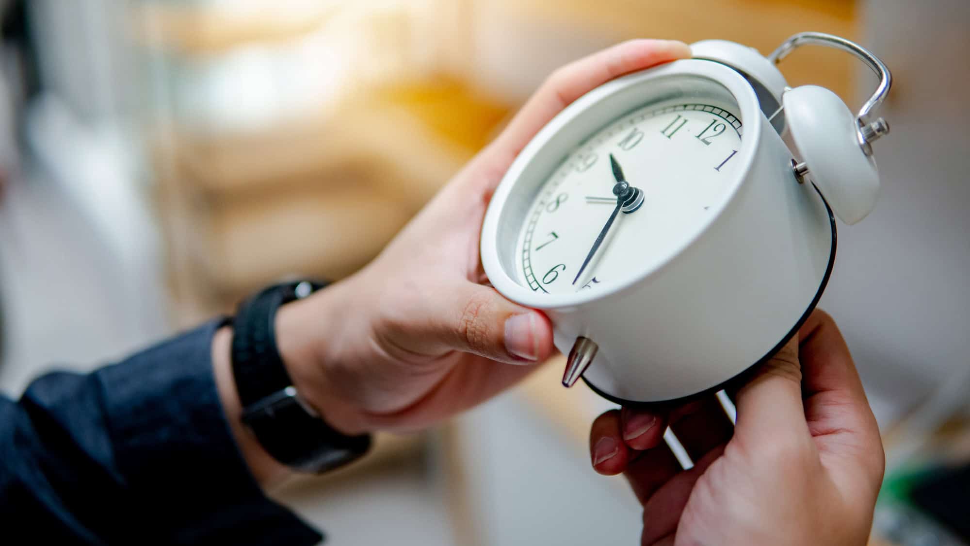 A close-up of a person adjusting a white vintage-style alarm clock with both hands. The person wears a dark-colored watch, and the blurred background suggests an indoor setting with warm lighting.