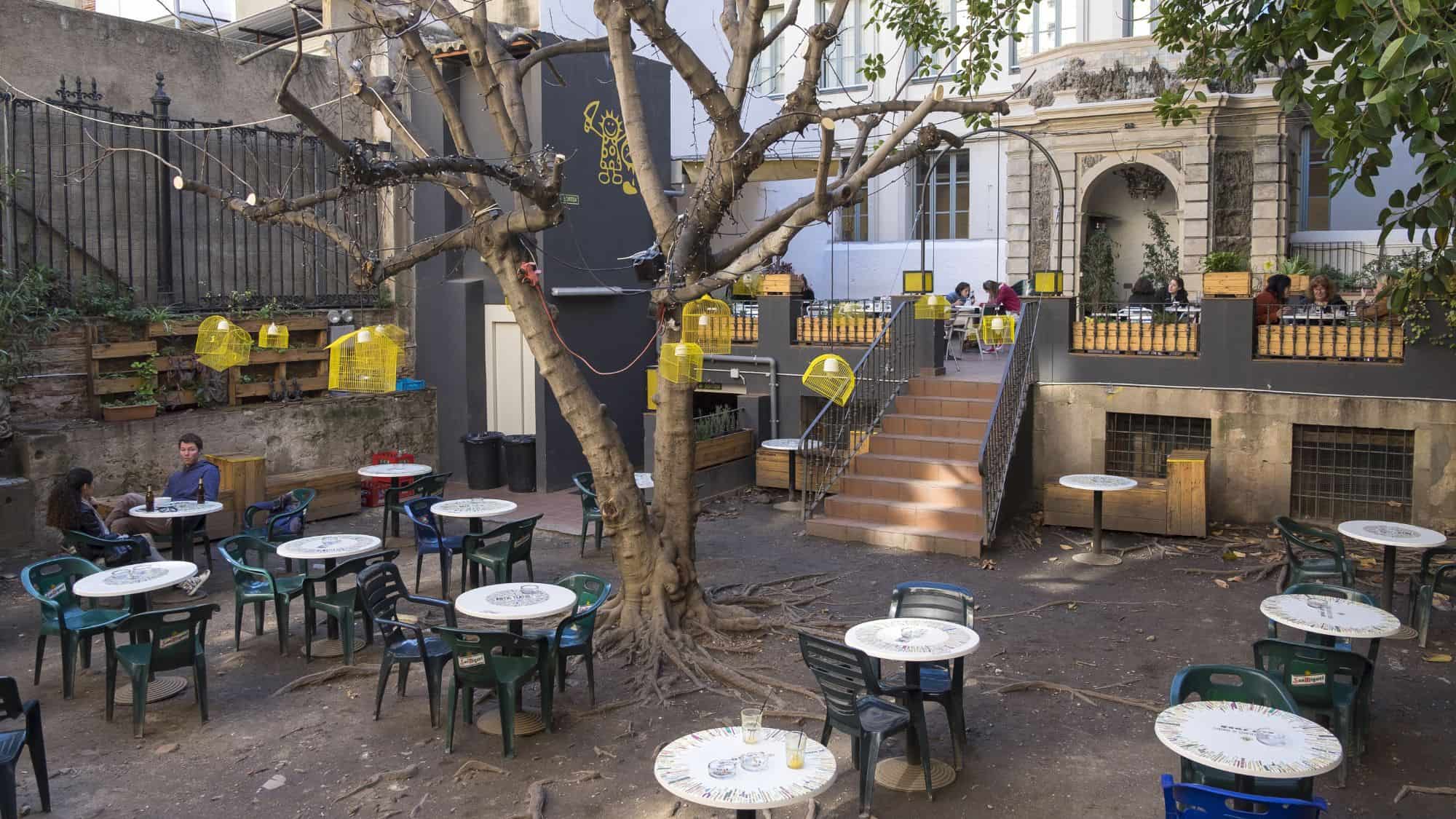 a courtyard with tables surrounding by old buildings. in the center of the courtyard sits a large shade tree.