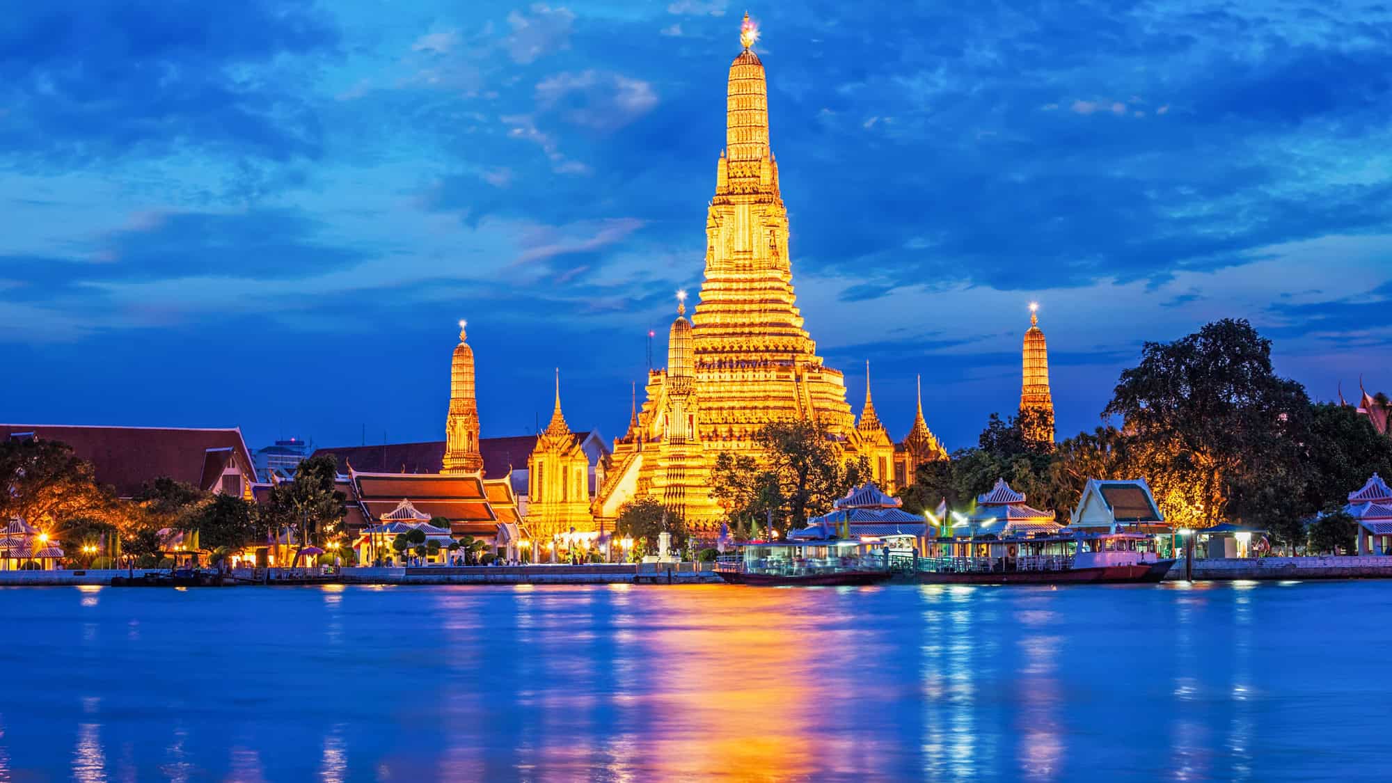 The iconic Wat Arun temple in Bangkok, glowing brightly with golden lights reflected in the calm waters of the Chao Phraya River, set against a deep blue evening sky.