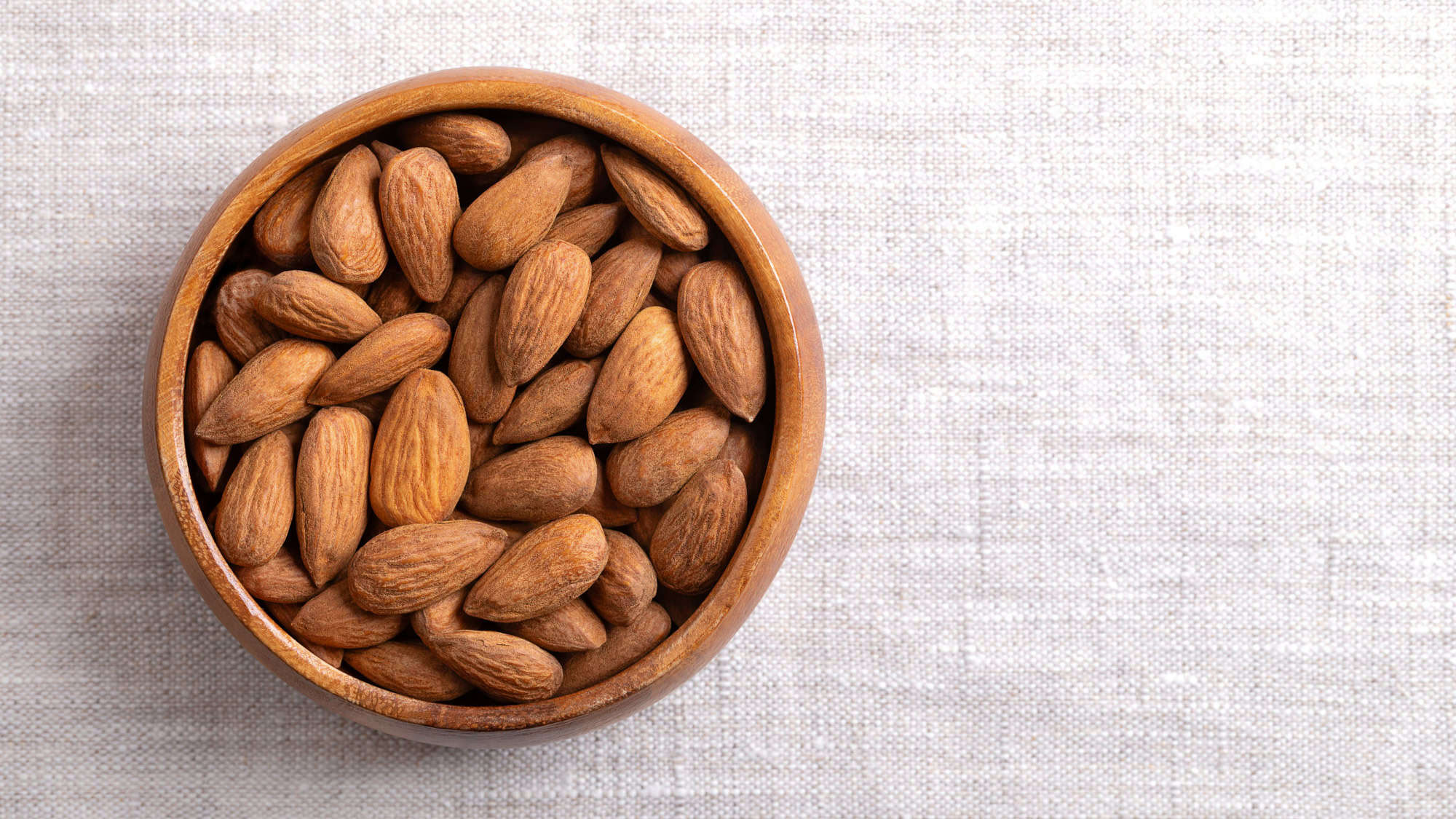 A wooden bowl filled with whole almonds sits on a neutral-colored fabric surface. The simple, earthy tones highlight the natural and healthy snack choice.
