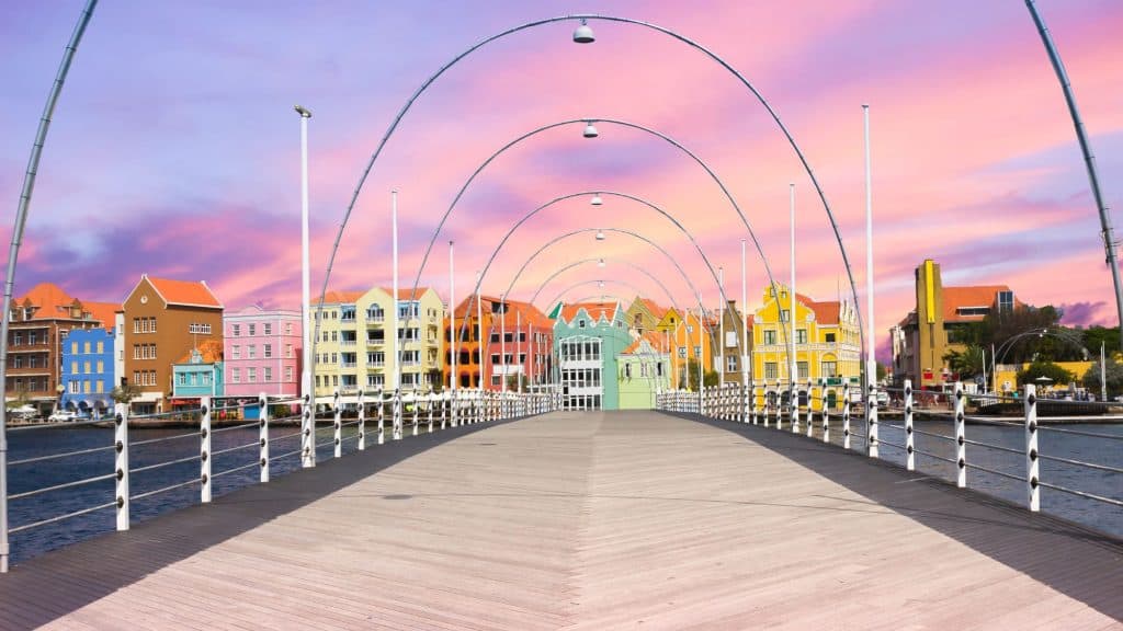 A vibrant waterfront cityscape viewed from a wooden pedestrian bridge with silver arches, featuring colorful buildings in shades of blue, orange, pink, and yellow, set against a pastel-hued sky at sunrise or sunset.