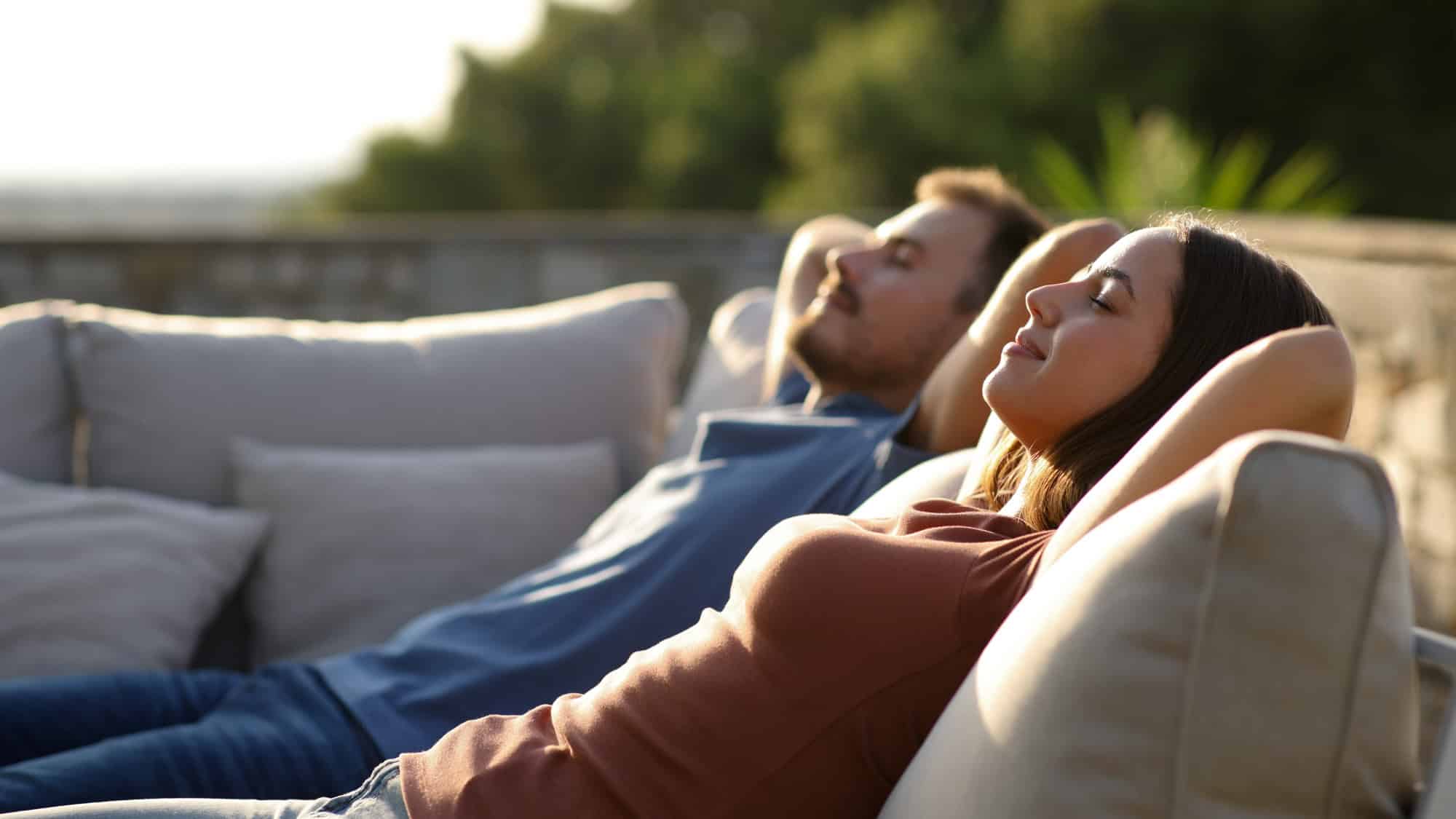 A relaxed couple lounges on a cozy outdoor couch, enjoying the fresh air. Both have their arms behind their heads, eyes closed, soaking in the peaceful atmosphere.