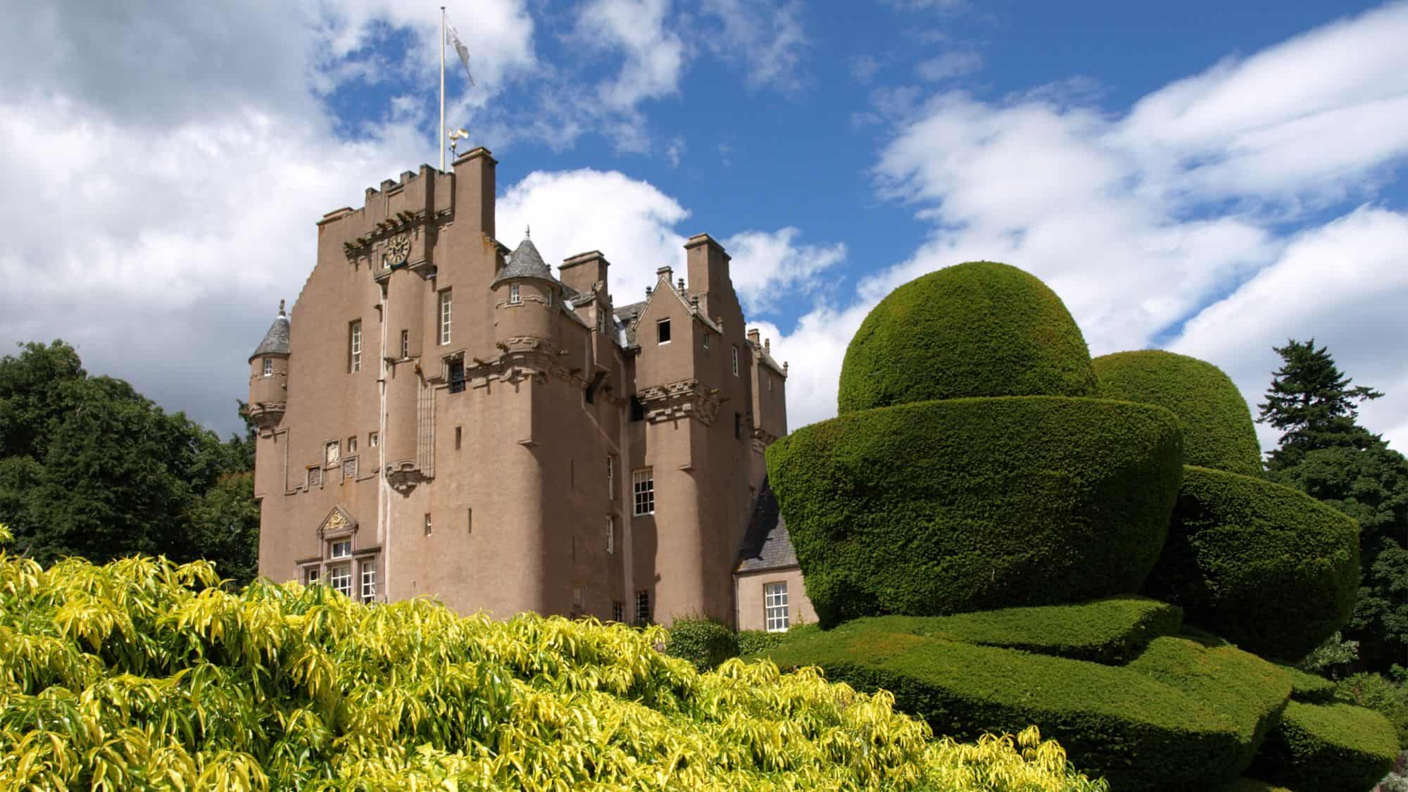 A sandstone castle with distinctive turrets and a clock tower, surrounded by trimmed hedges, green lawns, and a bright blue sky.