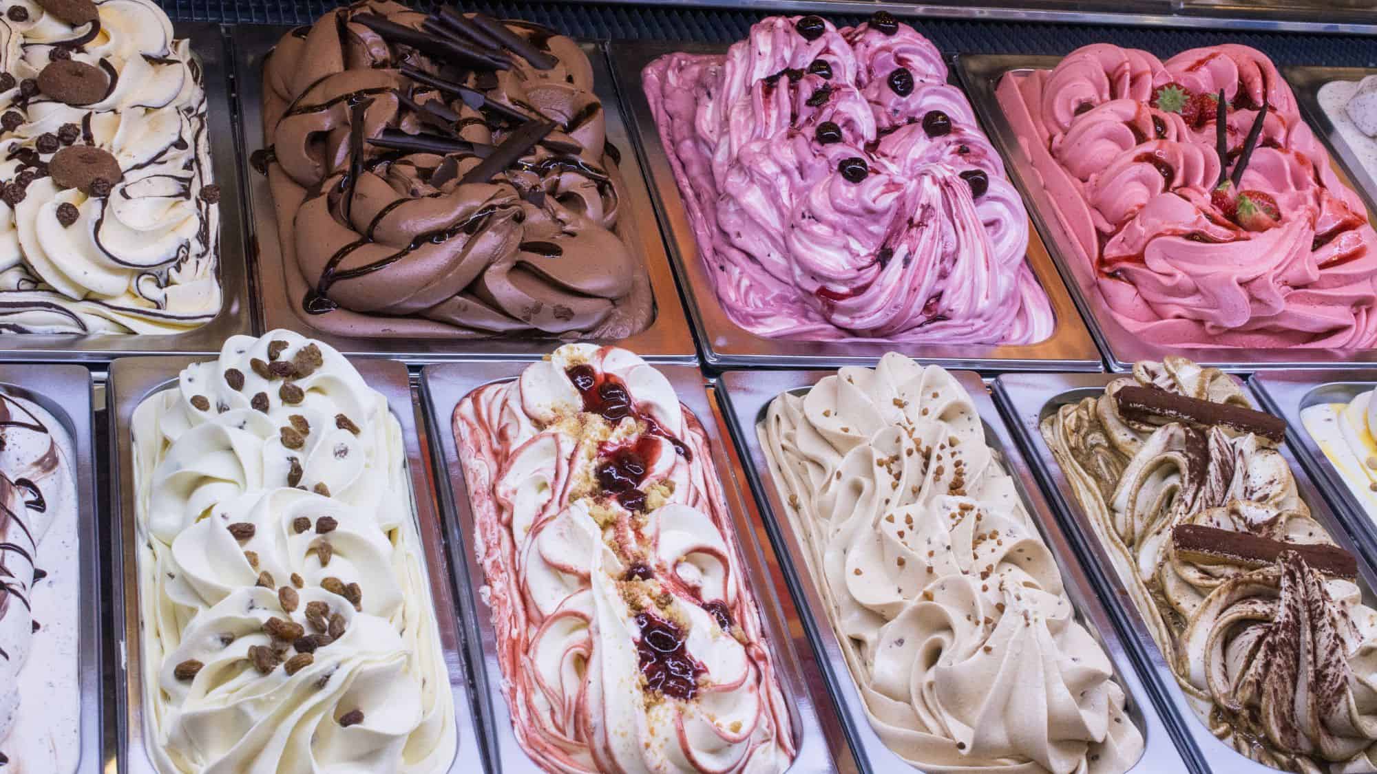 A vibrant selection of gelato flavors displayed in metal trays at a dessert shop, featuring a variety of colors and toppings such as chocolate drizzle, fruit garnishes, and swirls of sauce.