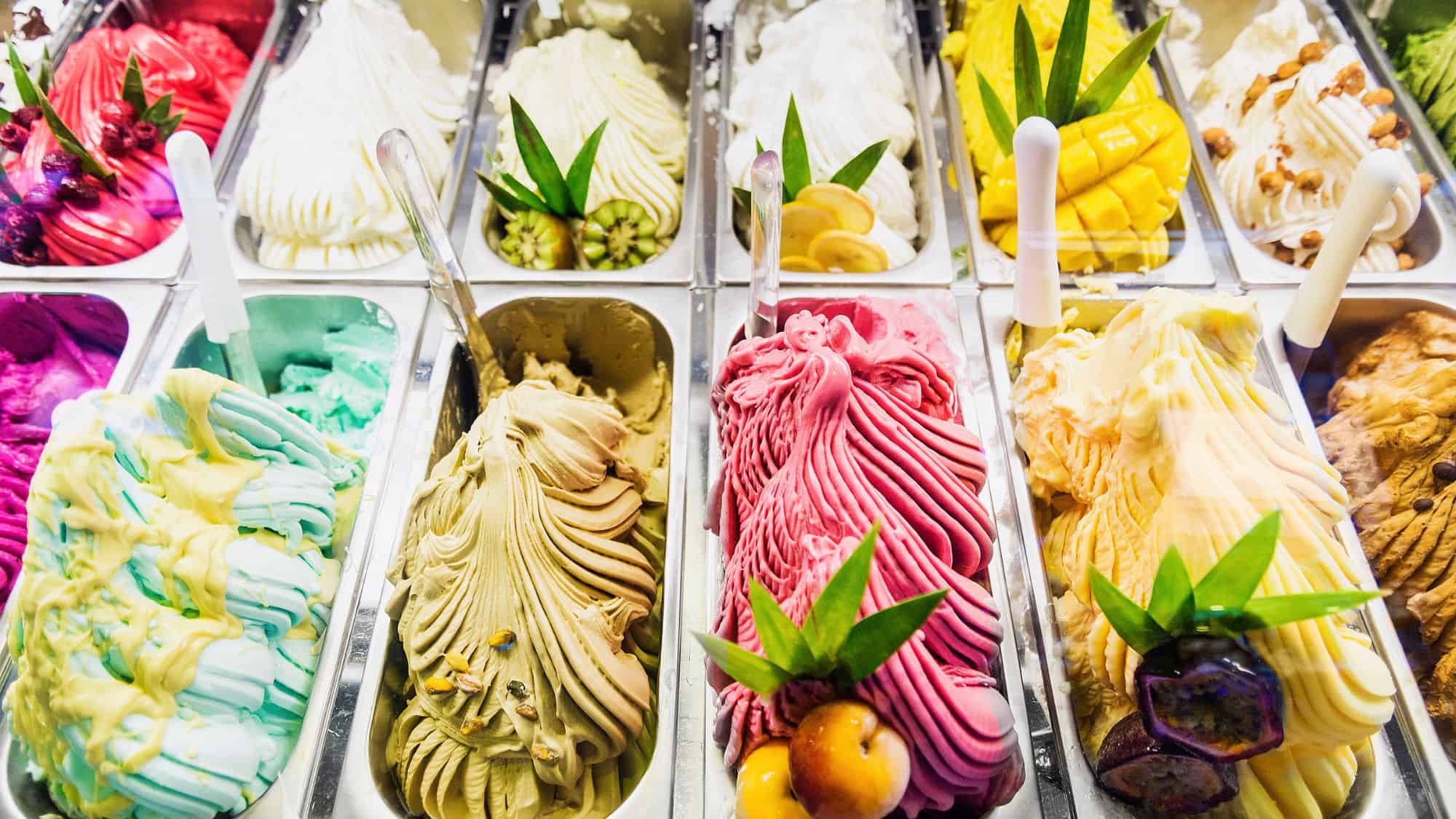 A colorful array of gelato flavors in a display case, each garnished with fresh fruit or toppings. The vibrant colors and intricate swirls make the gelato look especially appealing.