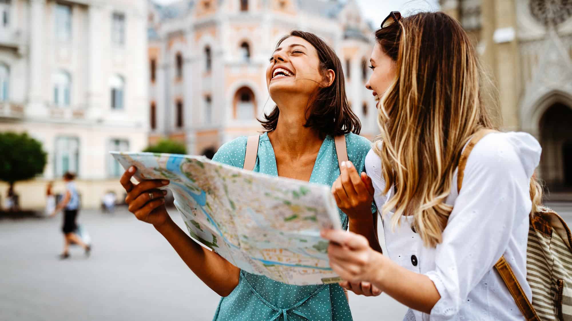 Two young women laugh and look at a city map while standing in a charming European-style plaza. Their excitement and casual outfits suggest they are tourists enjoying their trip.