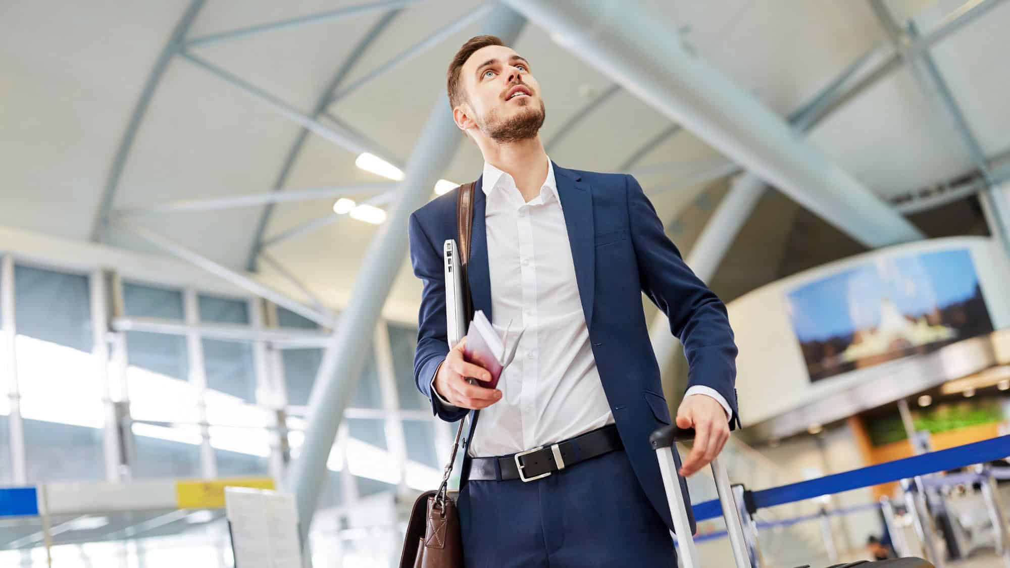 A professionally dressed man in a navy blue suit walks through an airport, holding a passport and rolling a suitcase. He looks upward as he navigates the terminal.