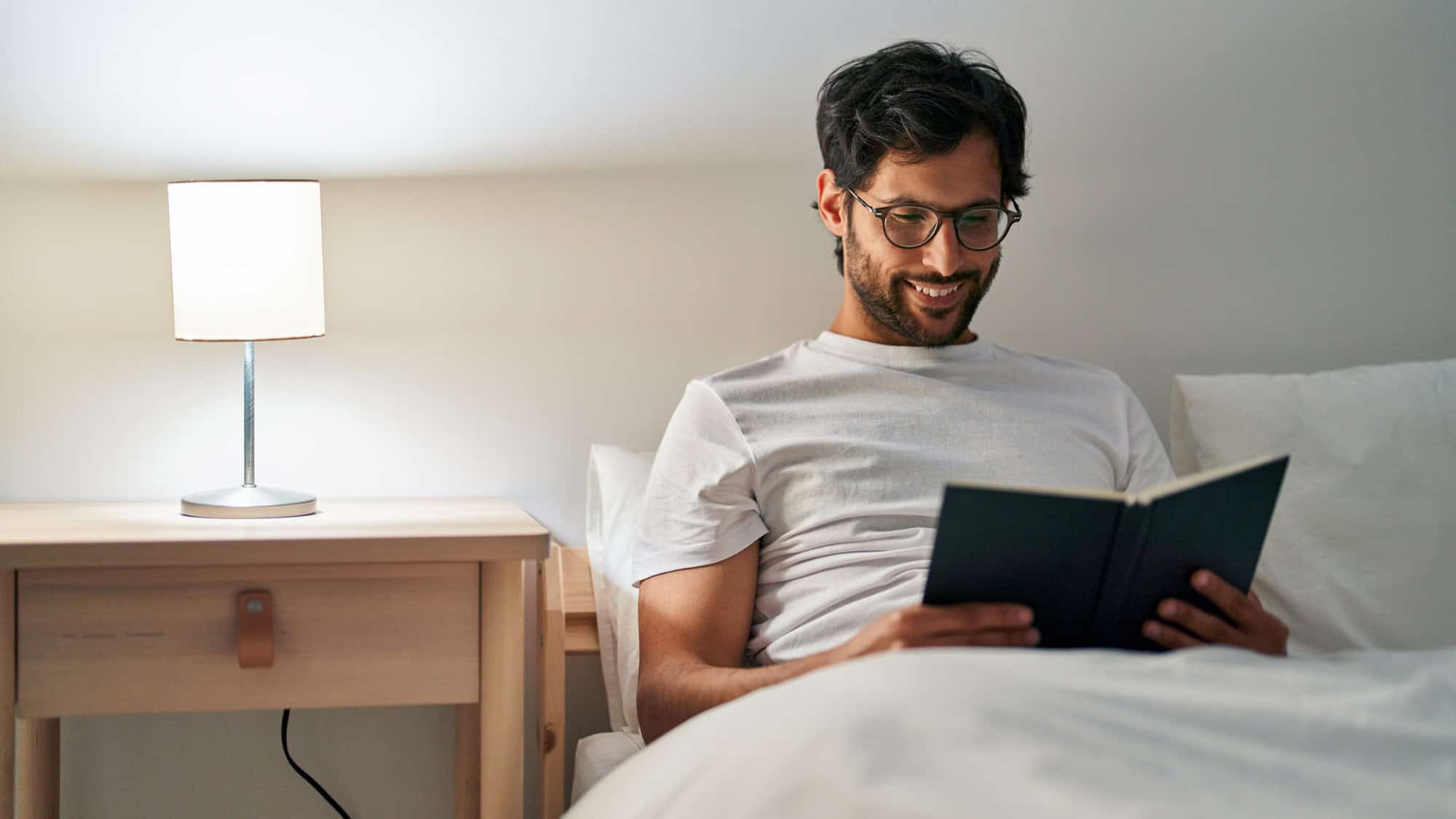 A man with glasses and a beard relaxes in bed, smiling while reading a book. A soft bedside lamp provides warm lighting, creating a peaceful nighttime routine.