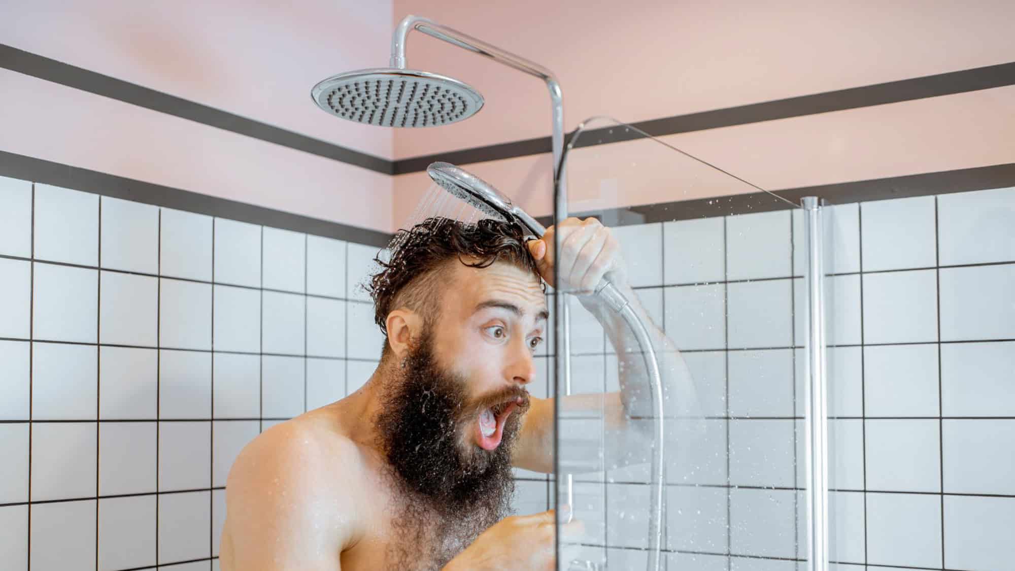 A bearded man in a shower reacts dramatically to an unexpected blast of cold water. His wide-eyed, open-mouthed expression adds humor to the moment.