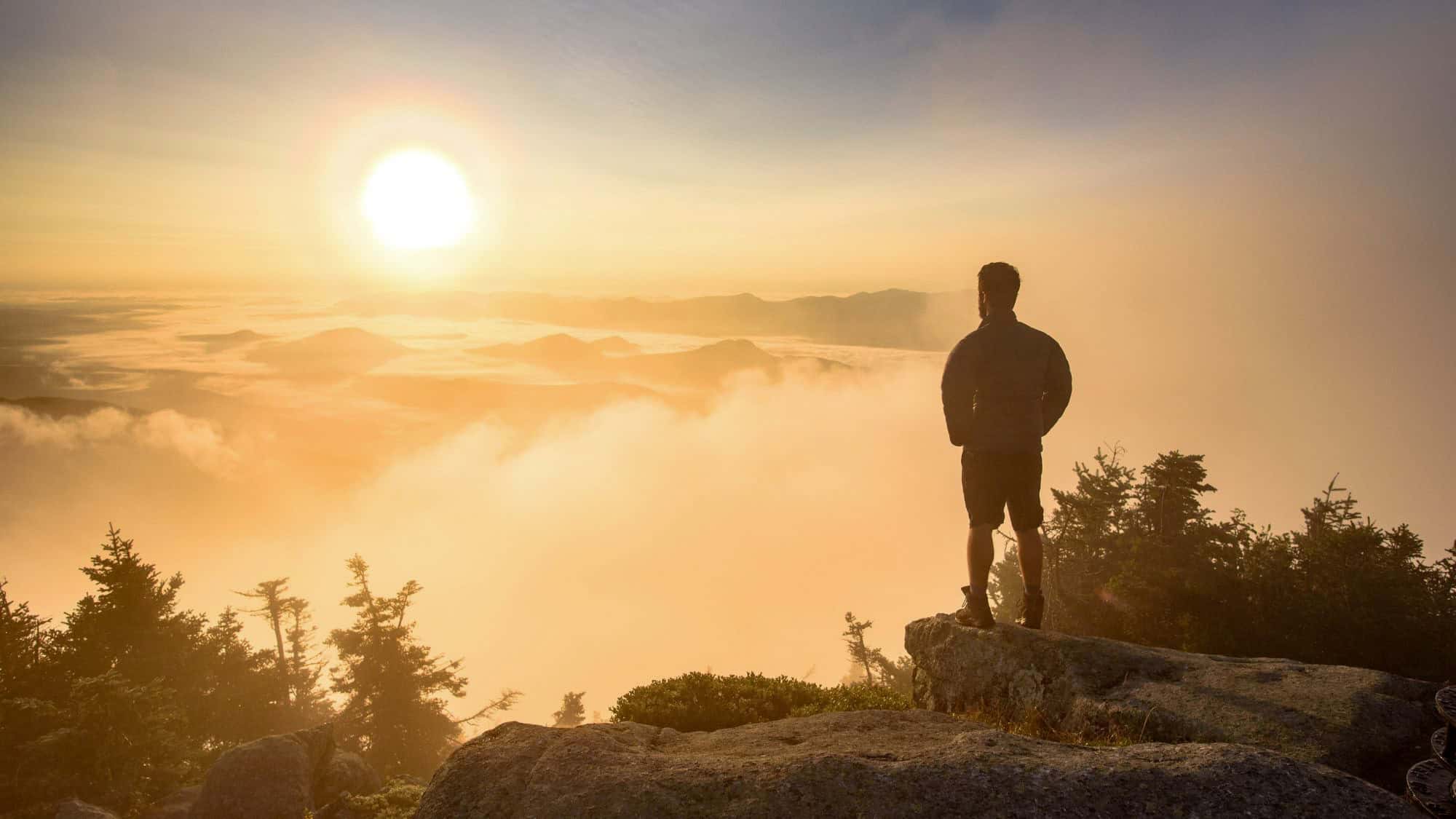 A silhouette of a man standing on a mountaintop, gazing at a golden sunrise over a vast landscape of clouds and rolling hills.