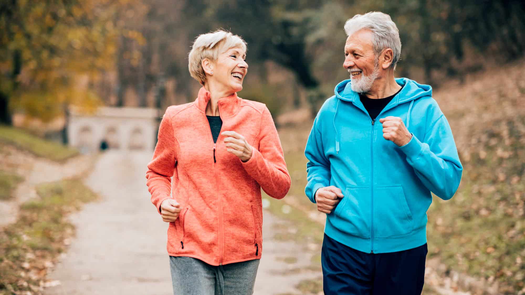 A happy elderly couple jogs together on a scenic outdoor path, dressed in sporty hoodies and activewear. Their smiles and laughter highlight the joy of staying active at any age.