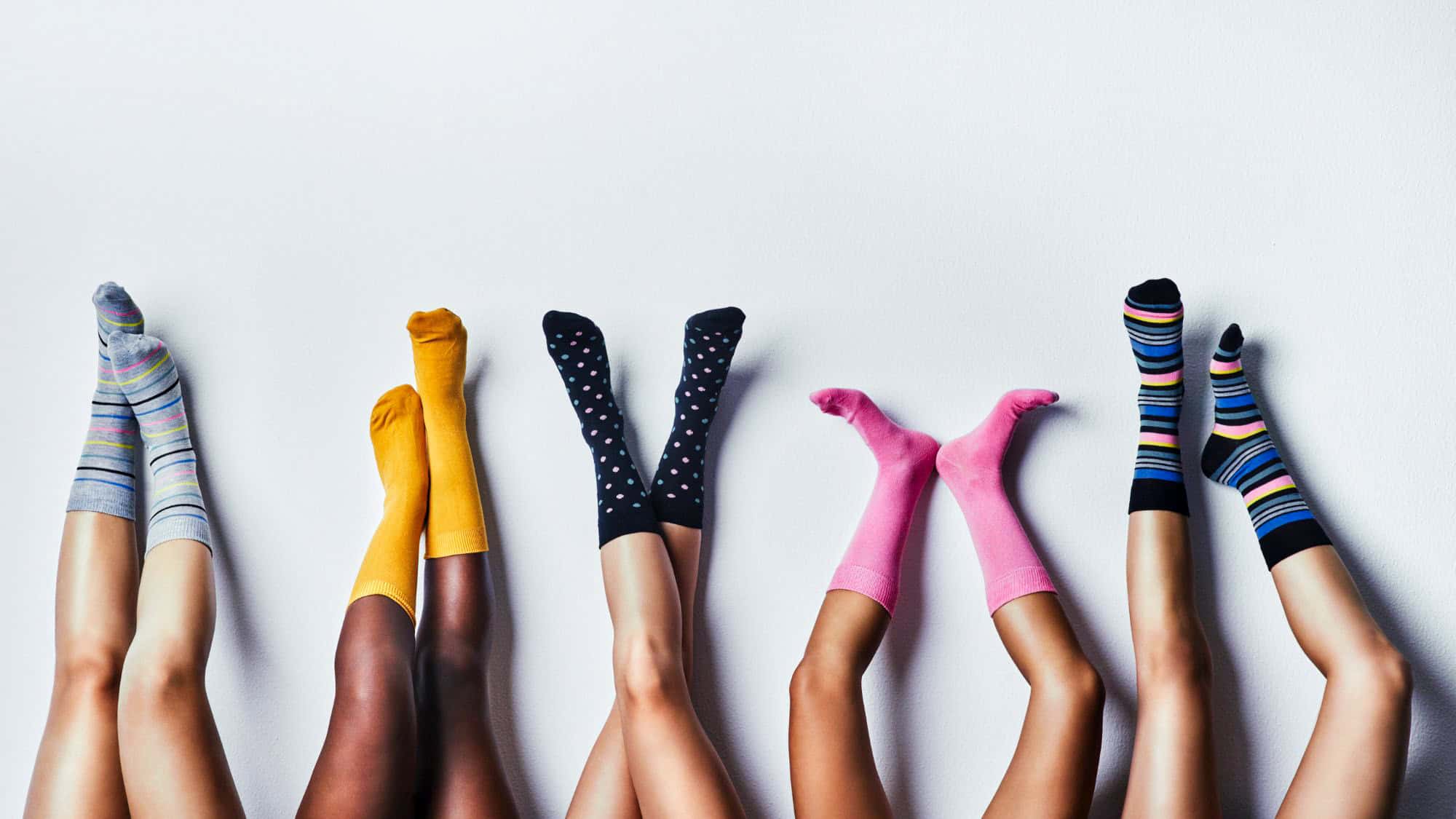 Several pairs of legs raised against a plain white wall, each wearing brightly colored and patterned socks. The mix of stripes, polka dots, and solid colors creates a playful and cozy vibe.