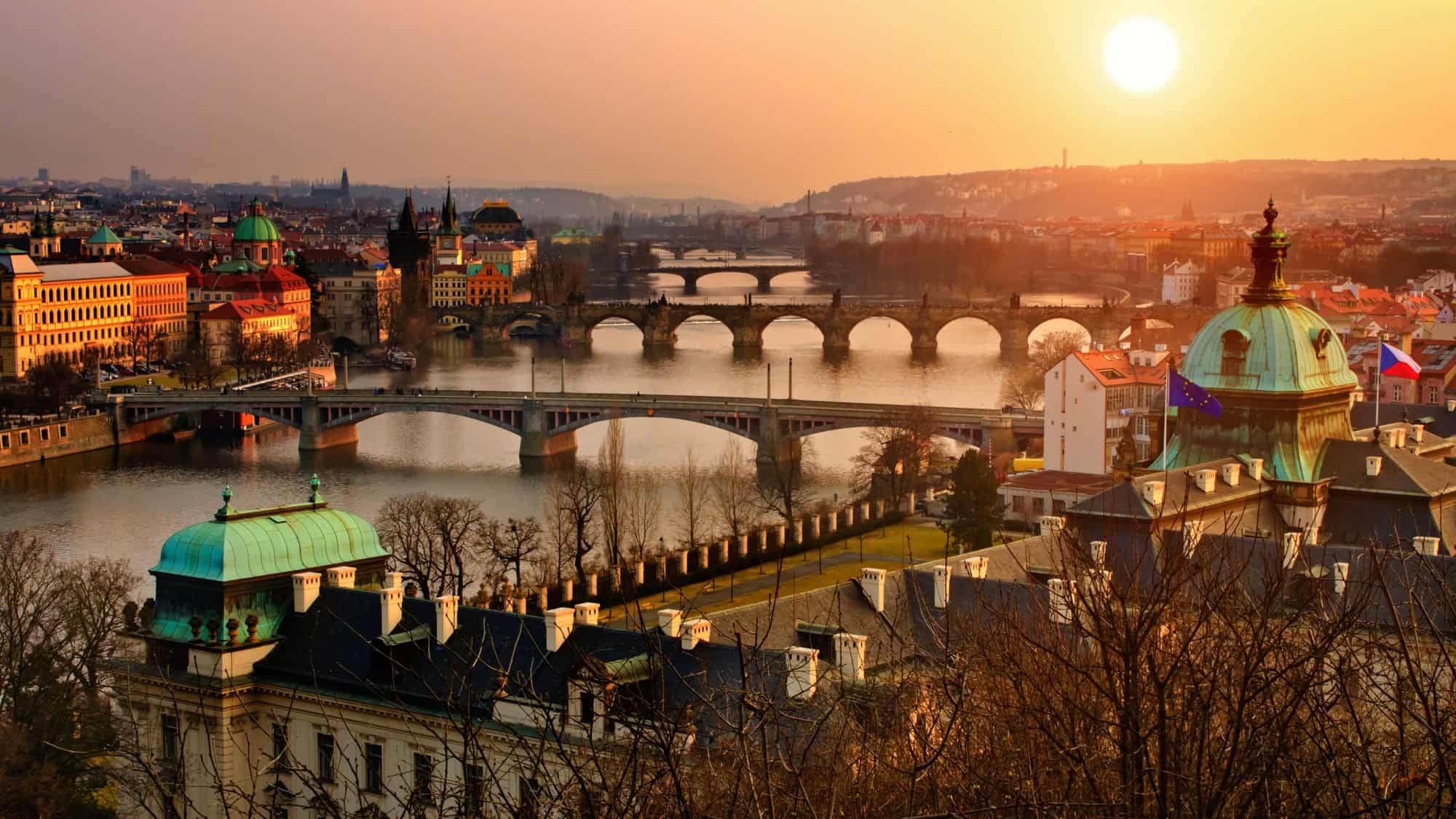 Multiple historic bridges span the Vltava River under a golden sunset in Prague, Czech Republic.