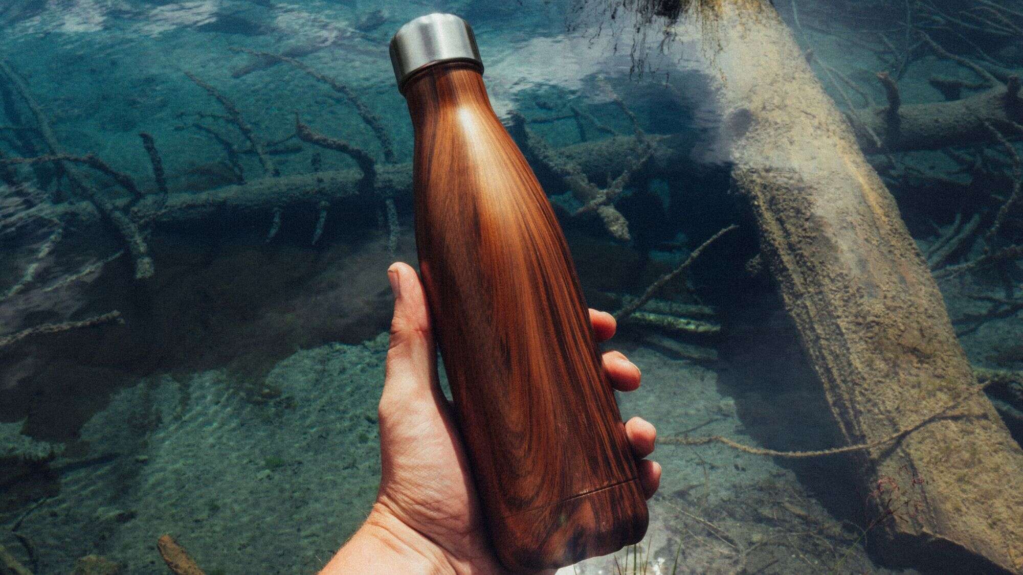 Hand holding a wood-patterned reusable water bottle against clear turquoise water with submerged logs.