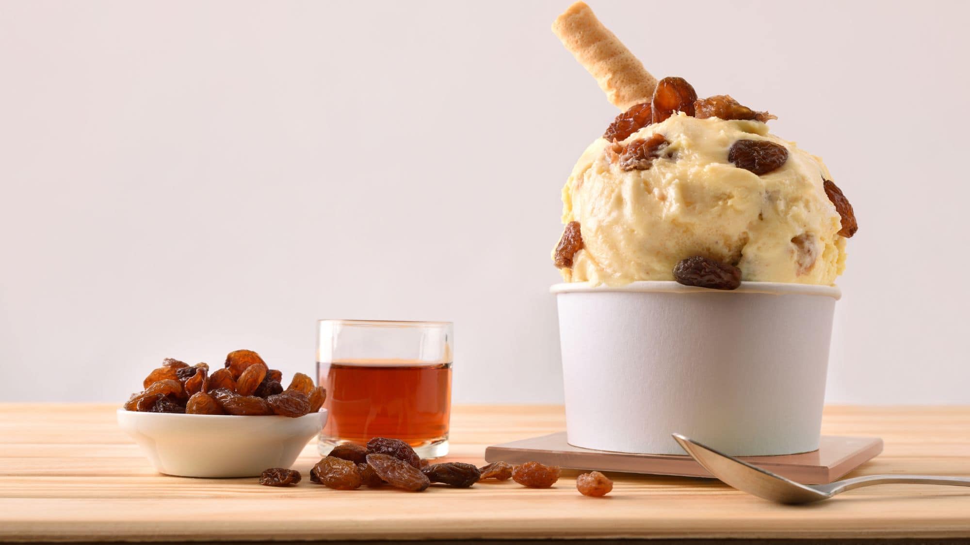 A scoop of rum and raisin ice cream served in a white cup, topped with plump raisins and accompanied by a small dish of raisins and a glass of rum on a wooden surface.