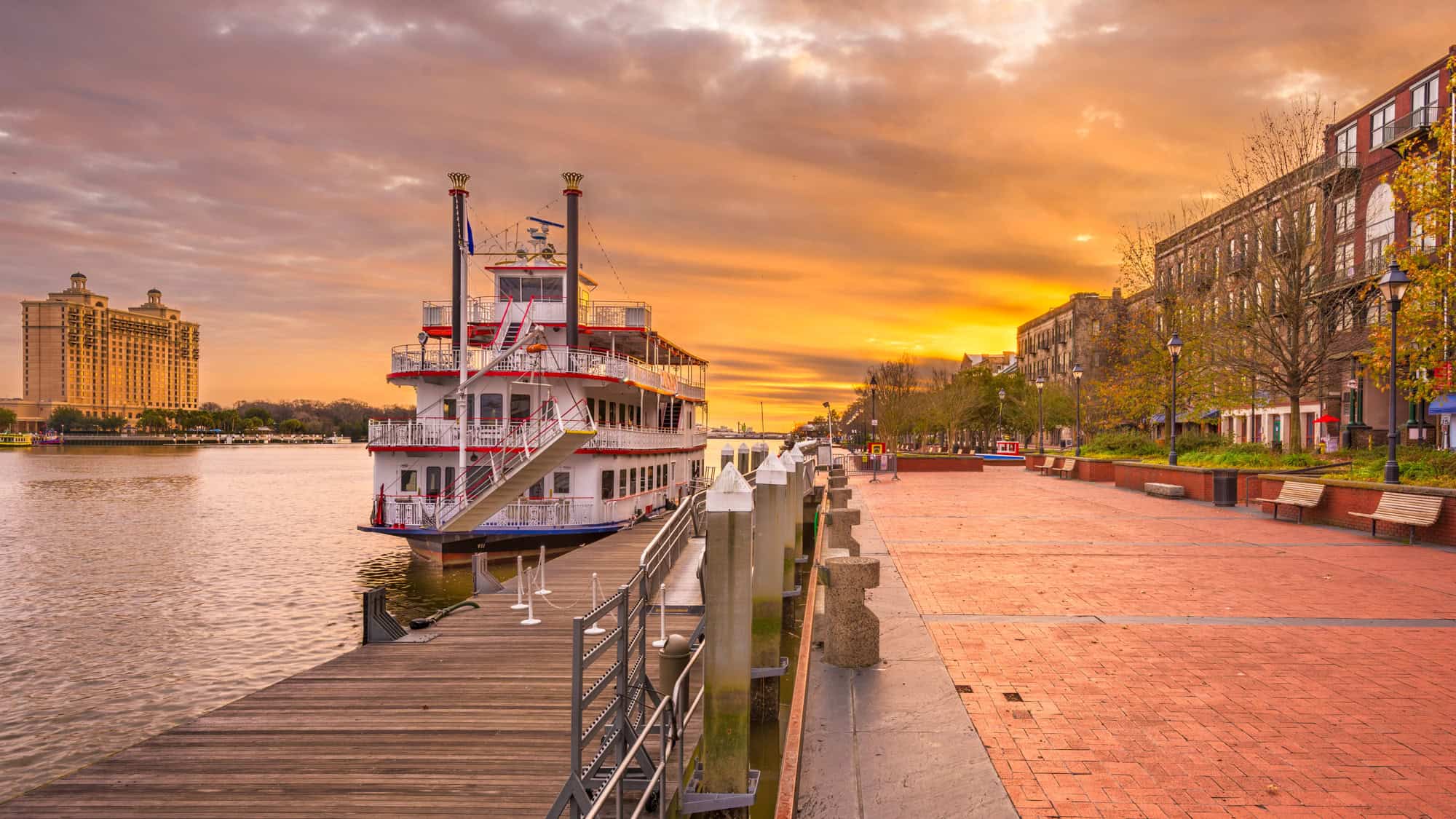 A classic riverboat is docked along the Savannah Riverwalk as the sky glows with golden and pink hues.