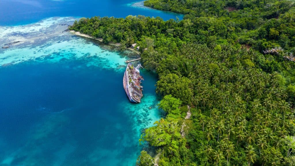 Aerial view of a tropical paradise featuring lush green islands surrounded by vibrant turquoise and deep blue waters, with a prominent rusted shipwreck nestled in a shallow cove near the shoreline.