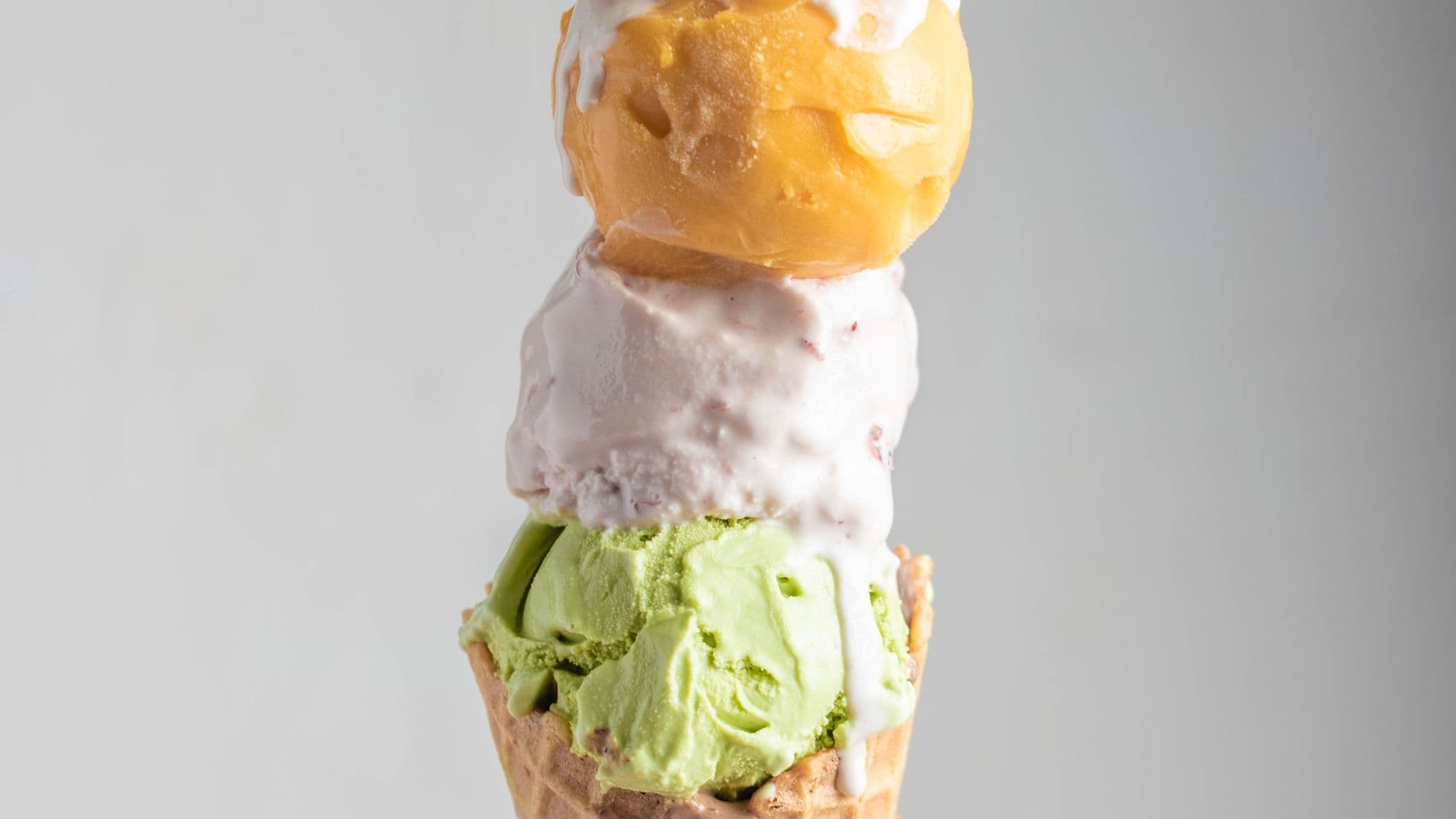 A tall ice cream cone with three scoops stacked on top of each other in different flavors, including pistachio green, creamy white, and mango yellow, set against a clean, white background.