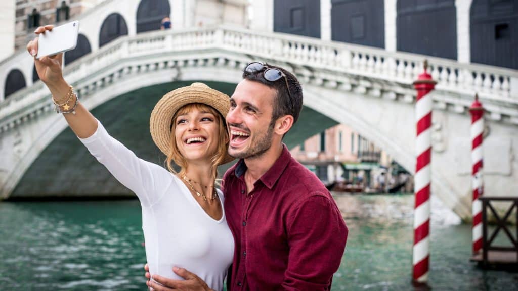 A smiling couple takes a selfie in front of the iconic Rialto Bridge over the Grand Canal. The bright day and historic architecture capture the charm of the city.