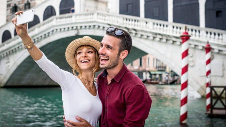 A smiling couple takes a selfie in front of the iconic Rialto Bridge over the Grand Canal. The bright day and historic architecture capture the charm of the city.