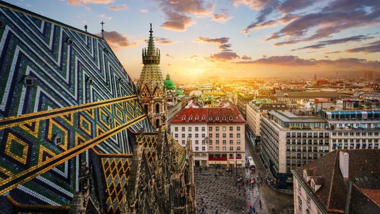 An iconic view of Vienna showcasing the ornate, geometric tiled roof of St. Stephen’s Cathedral at sunset. The cityscape spreads out in the background with historic and modern buildings under a colorful sky.