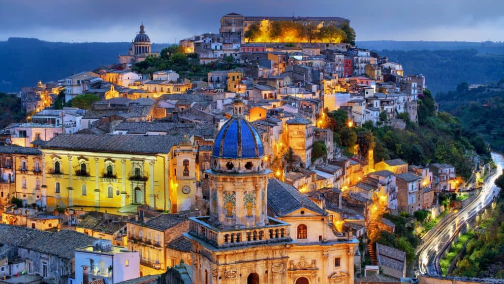 A picturesque hillside town in Sicily illuminated at dusk, featuring narrow winding streets and a mix of historic buildings with warm-toned facades. The focal point is a church with a striking blue-tiled dome, surrounded by traditional architecture and lush greenery.