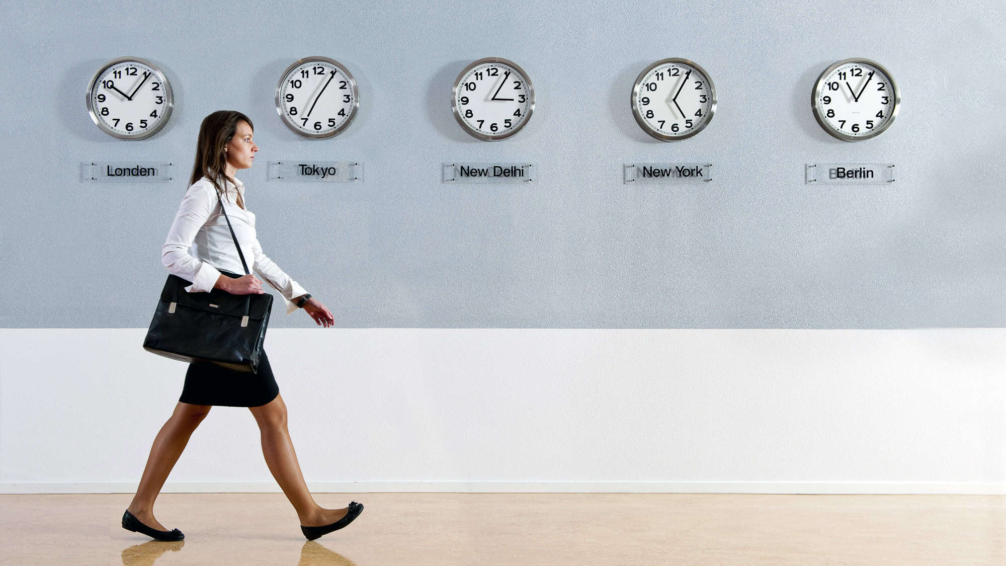 A businesswoman in a white blouse and black skirt walks past a wall displaying multiple clocks labeled with different world cities. The clocks indicate various time zones, emphasizing an international setting.