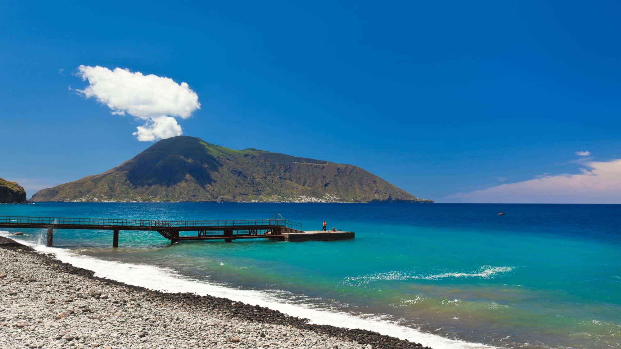 A scenic beach on the Aeolian Islands, Sicily, with black pebbles, clear turquoise waters, and a wooden pier extending into the sea. A green volcanic mountain rises in the background under a bright blue sky.