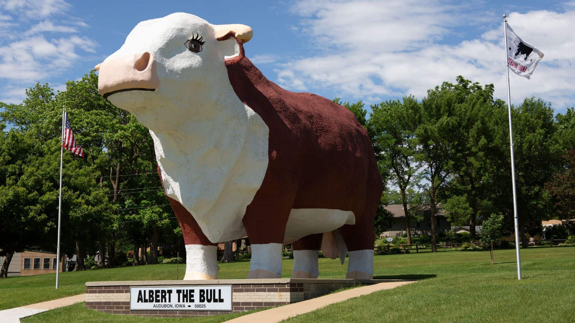 A massive statue of Albert the Bull, a giant Hereford cow, stands on a green lawn with an American flag flying nearby. A sign below reads "Albert the Bull, Audubon, Iowa," marking this as the world’s largest bull statue.