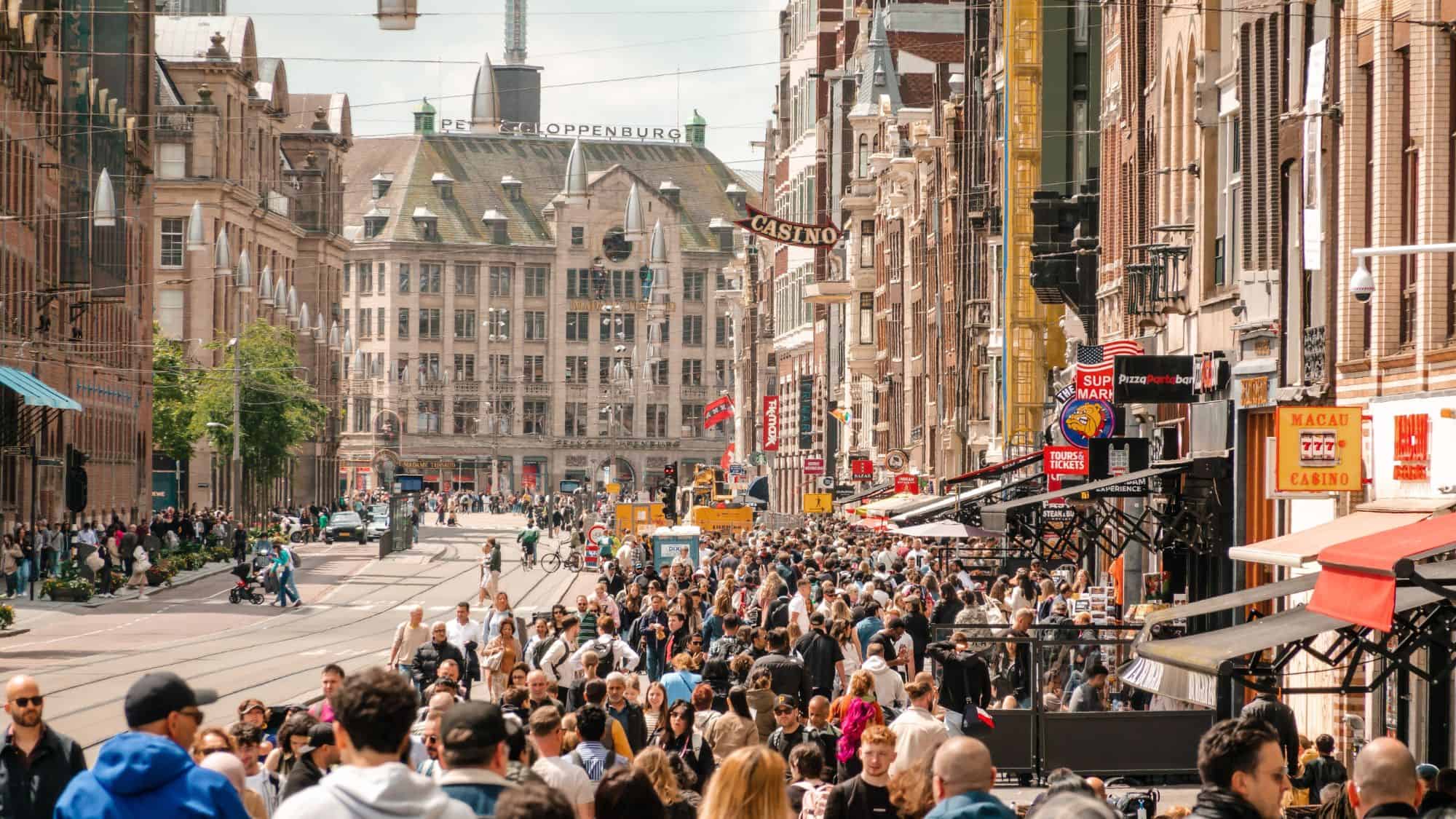 A crowded shopping street in Amsterdam, Netherlands, filled with people walking past historic European architecture and modern storefronts. The tram tracks run through the center, with lively cafés, casino signs, and businesses on both sides.
