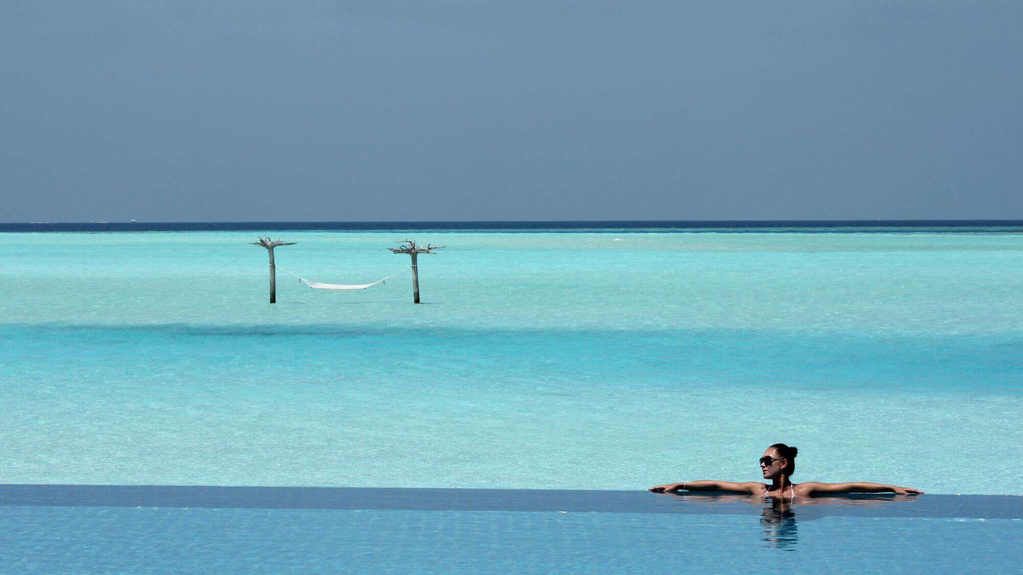 A woman relaxes in an infinity pool overlooking the ocean, with a hammock suspended between two wooden posts in the water. The horizon stretches endlessly, merging sea and sky.