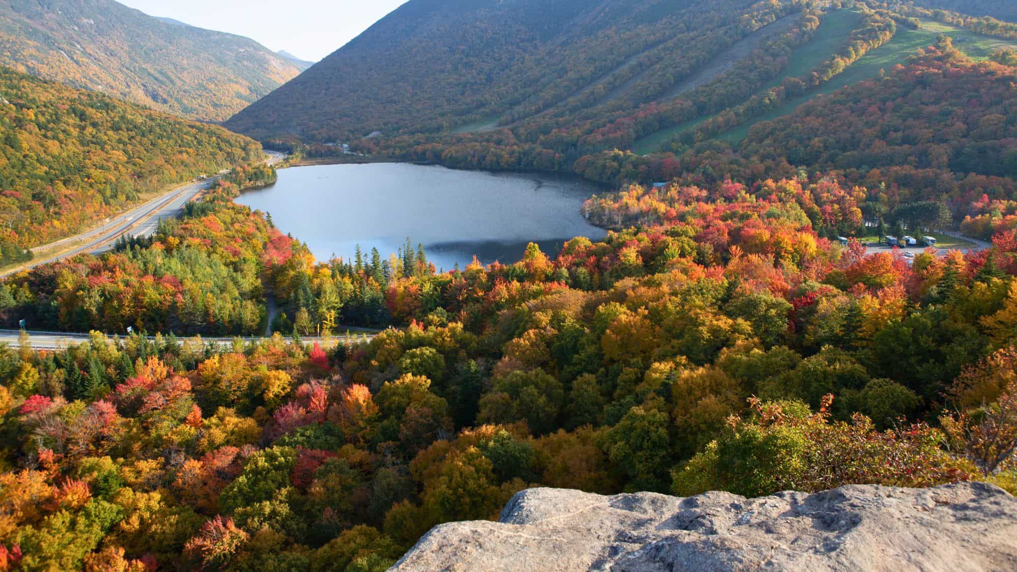 An aerial view of a serene lake nestled between forested mountains, with vibrant fall foliage in shades of orange, red, and yellow. A winding road traces the shoreline, highlighting the natural beauty of the lookout.