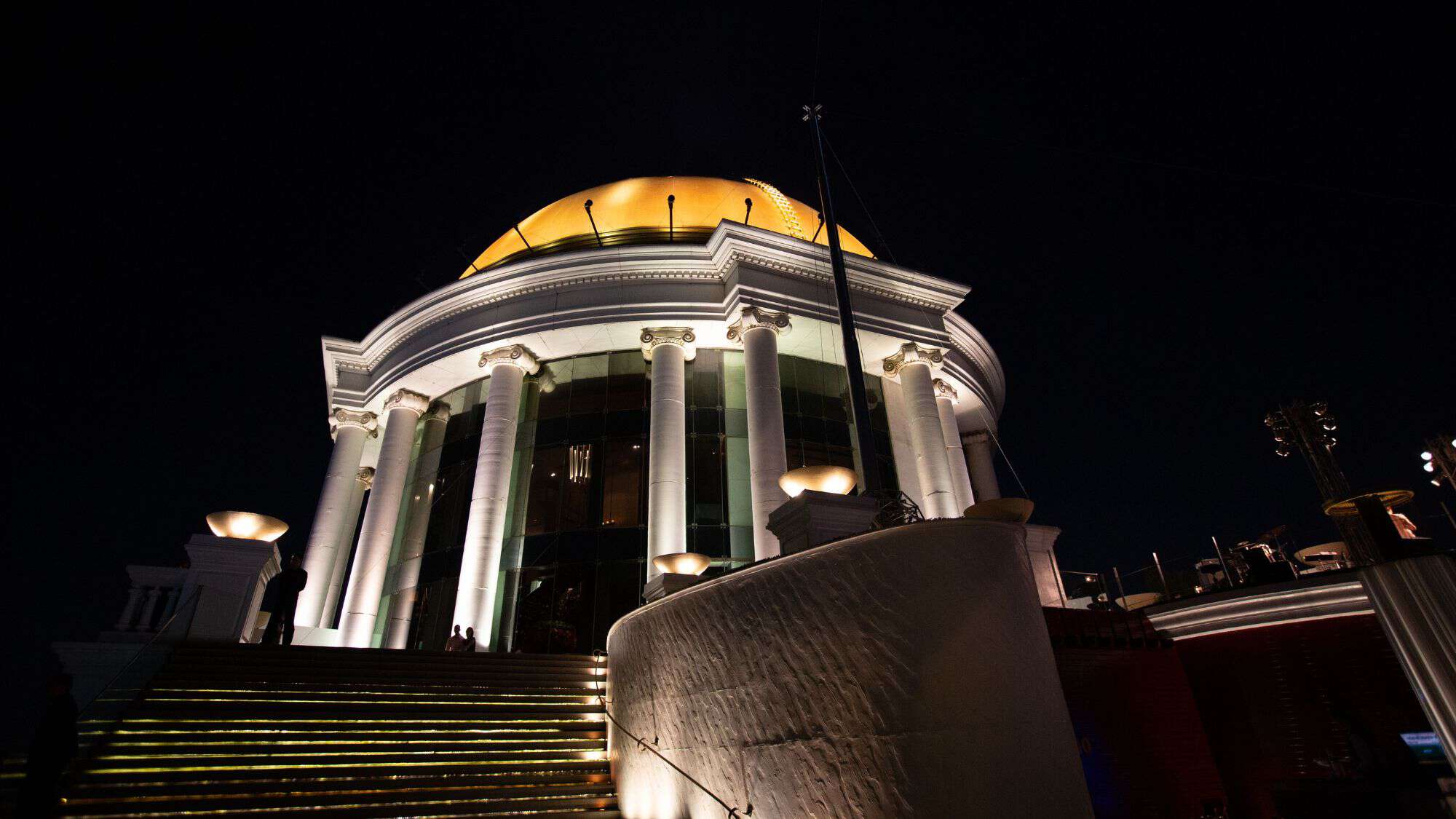 A grand, illuminated rooftop bar in Bangkok at night, featuring towering white columns and a golden dome. The steps leading up to the entrance glow with golden lighting, creating a luxurious ambiance against the dark sky.