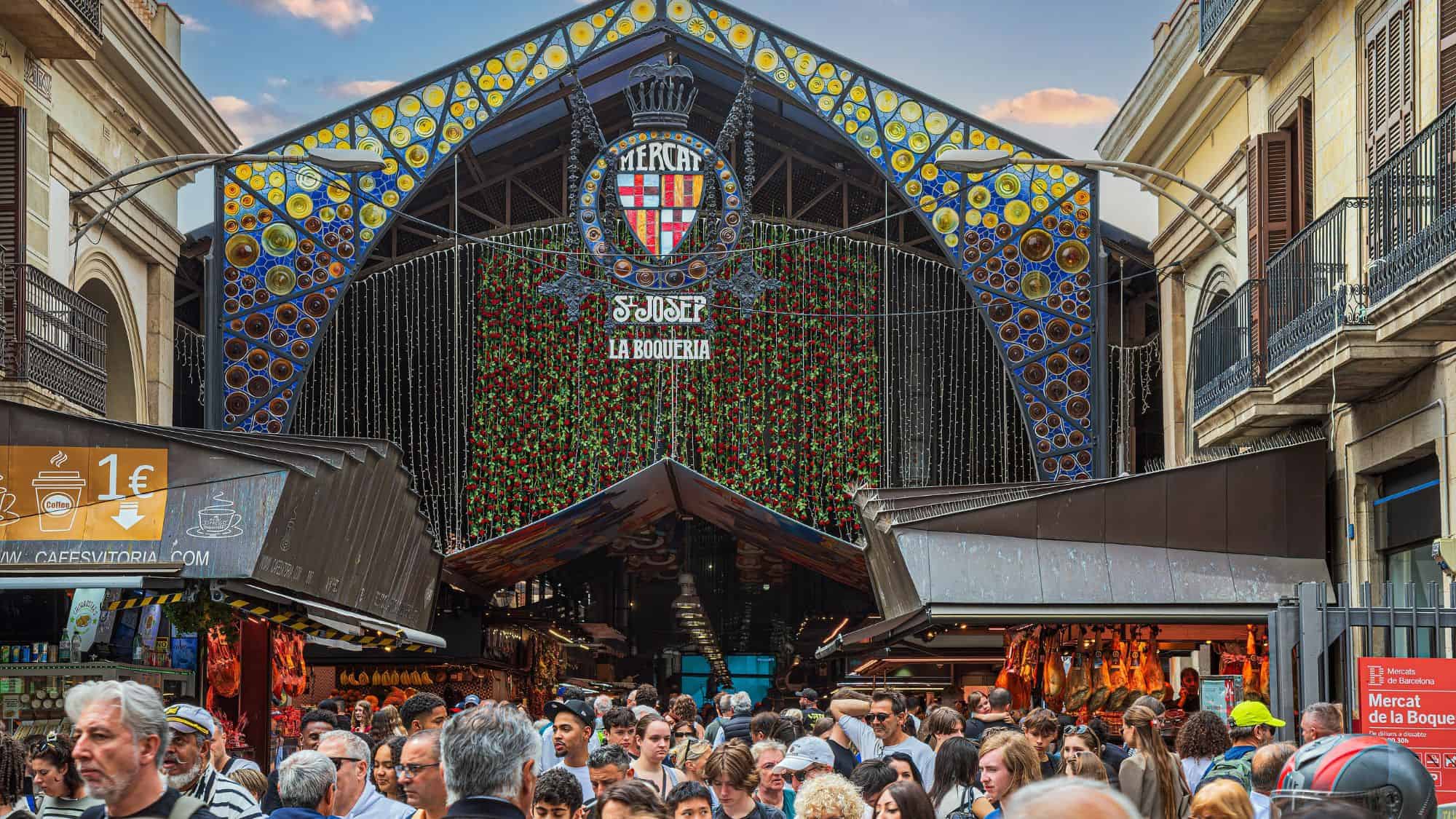 The entrance to La Boqueria Market in Barcelona, Spain, adorned with colorful glass mosaics and the market’s emblem. A dense crowd of shoppers and tourists fills the space, exploring fresh produce, meats, and local delicacies.