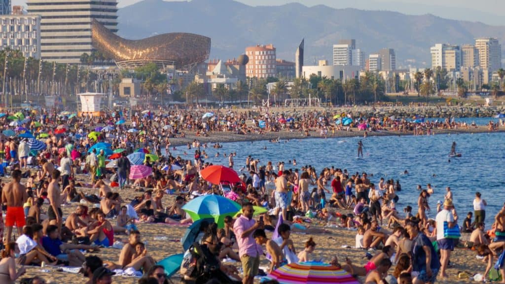 A lively and crowded beach in Barcelona, Spain, filled with sunbathers, colorful umbrellas, and beachgoers enjoying the Mediterranean shoreline. The cityscape, including the iconic golden fish sculpture by Frank Gehry, is visible in the background.