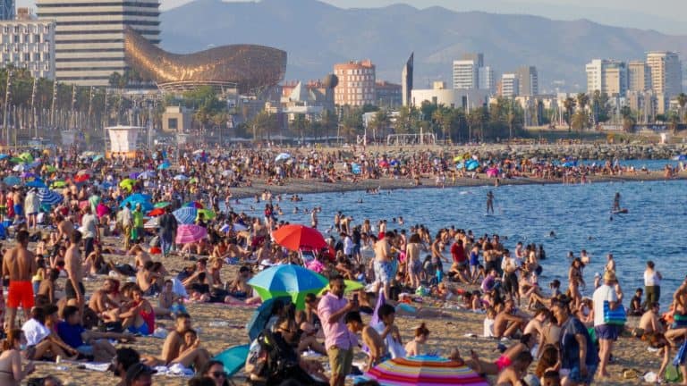 A lively and crowded beach in Barcelona, Spain, filled with sunbathers, colorful umbrellas, and beachgoers enjoying the Mediterranean shoreline. The cityscape, including the iconic golden fish sculpture by Frank Gehry, is visible in the background.
