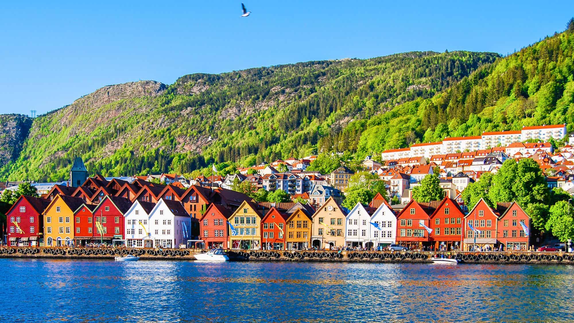 A row of traditional Norwegian wooden houses painted in bright colors along the harbor. The backdrop of lush green hills and a deep blue fjord makes the scene both lively and scenic.