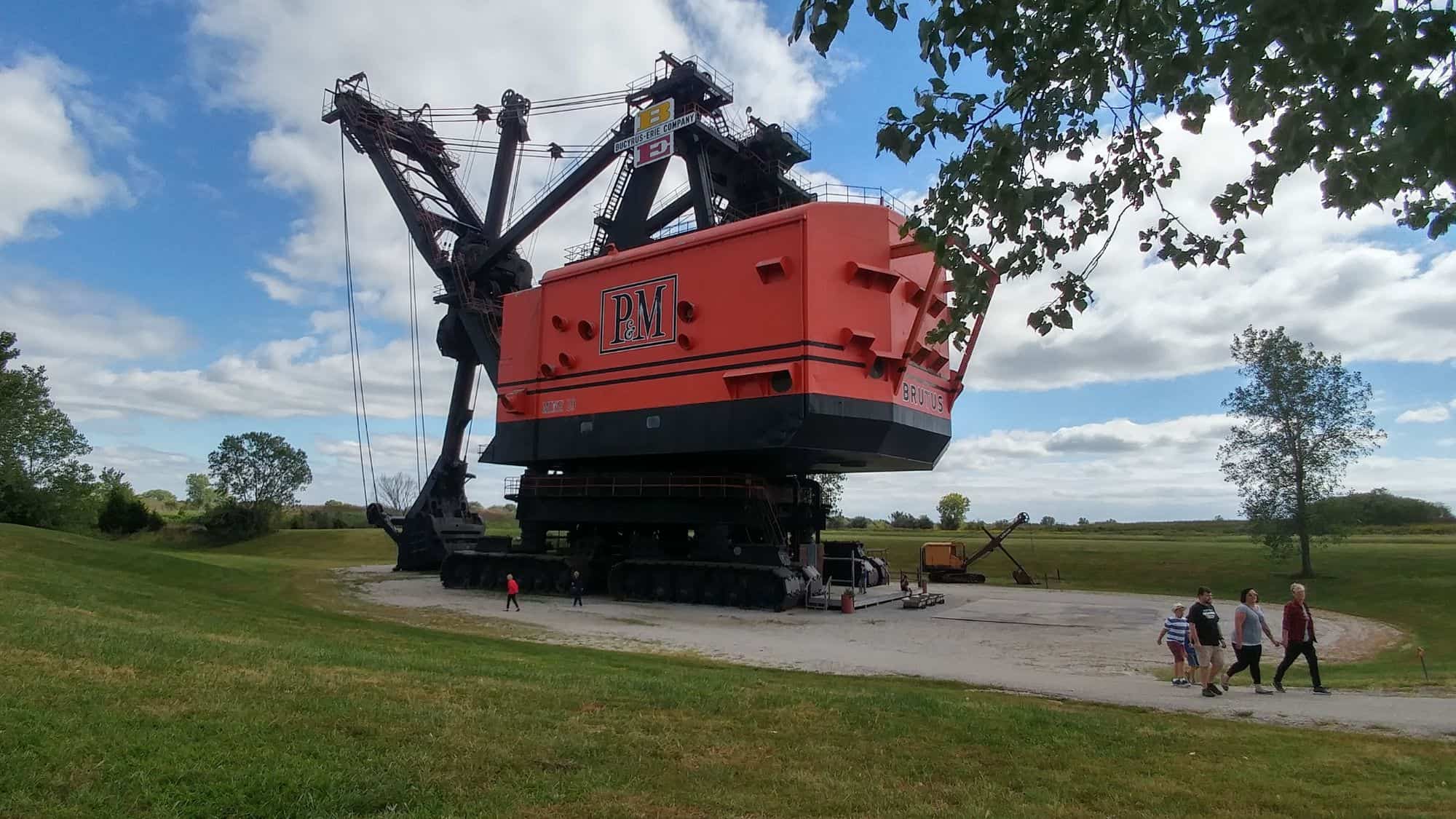 A massive, retired electric shovel painted in bright orange and black, towering over visitors at an outdoor exhibit. The machine, once used for strip mining, is displayed against a backdrop of green grass and blue skies.