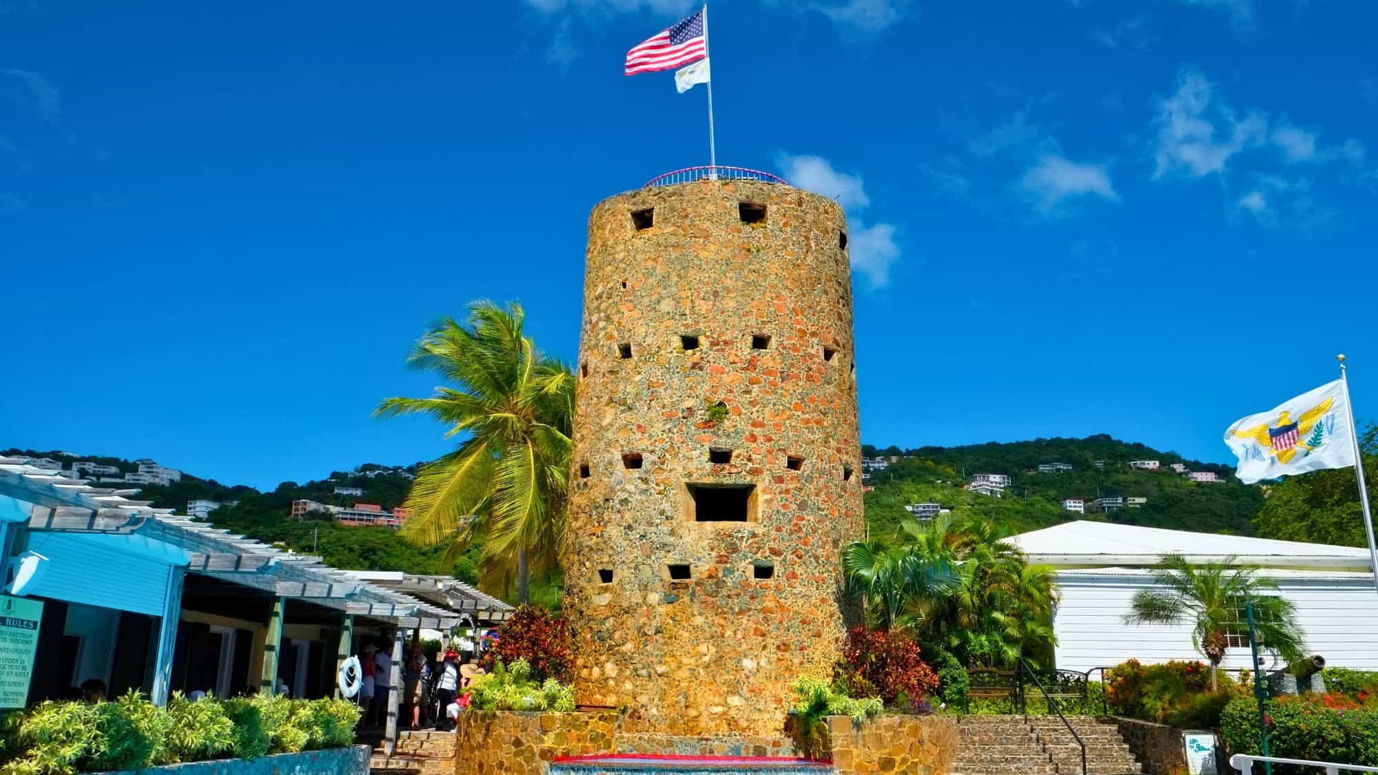 A historic round stone tower stands tall with American and U.S. Virgin Islands flags flying above. The tower is surrounded by tropical plants, a palm tree, and colorful flowers, with a clear blue sky overhead.