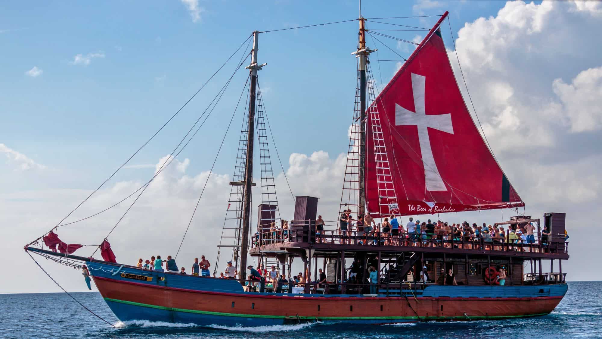 A large wooden pirate-style party boat with a red sail featuring a white cross cruises through the blue waters of Bridgetown, Barbados, filled with people enjoying the sunny day.