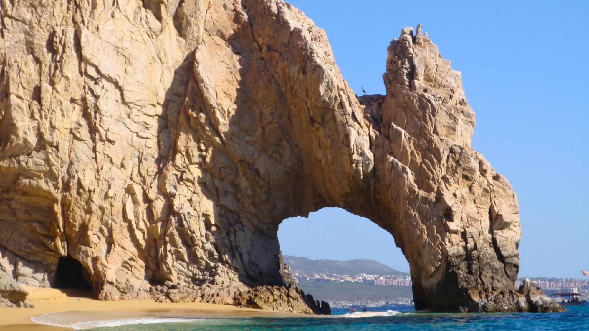 A stunning natural rock formation known as El Arco in Cabo San Lucas, Mexico. The massive stone arch rises above the ocean, with a sandy beach below and the city skyline visible in the background.