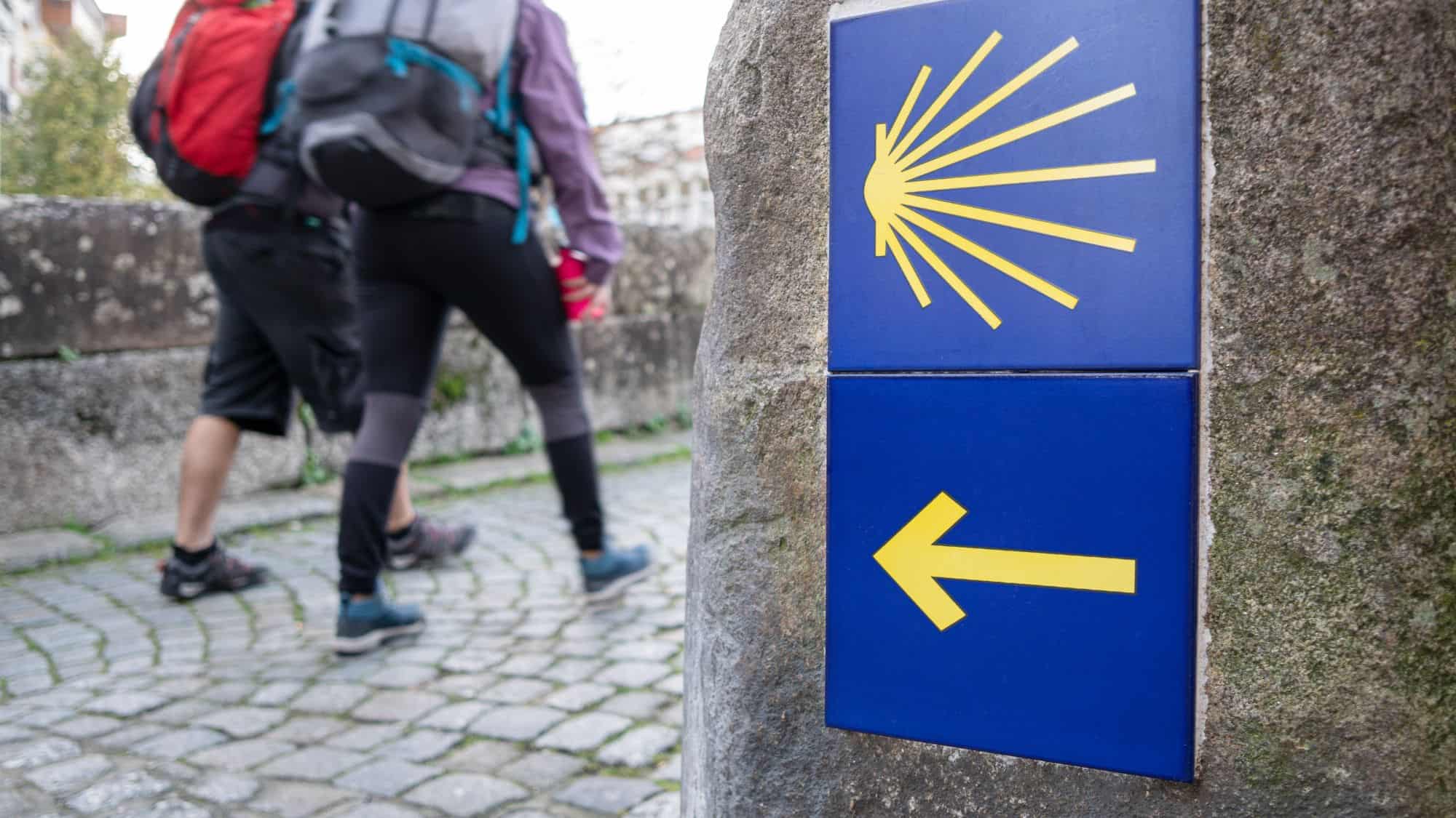 Two pilgrims walking along the cobblestone path of the famous Camino de Santiago, with a stone marker in the foreground displaying the iconic yellow scallop shell and arrow symbol on a blue background.