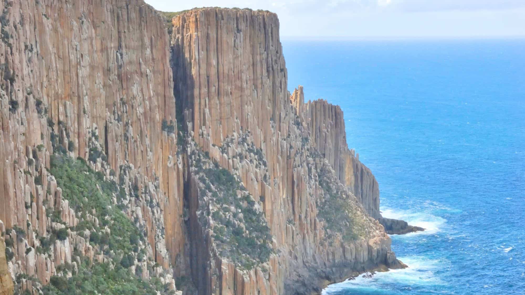 Dramatic cliffs rise sharply from the deep blue sea, with vertical rock formations creating a rugged coastline. Green vegetation clings to the edges, highlighting the stark contrast between land and ocean.