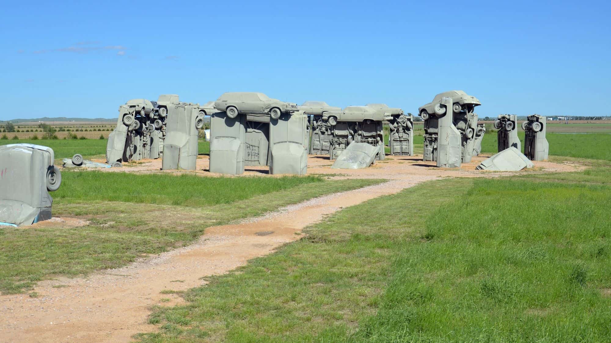 A roadside attraction featuring "Carhenge," a replica of England's Stonehenge made entirely of stacked and half-buried vintage cars, painted in a uniform gray. The installation stands in an open grassy field under a bright blue sky.