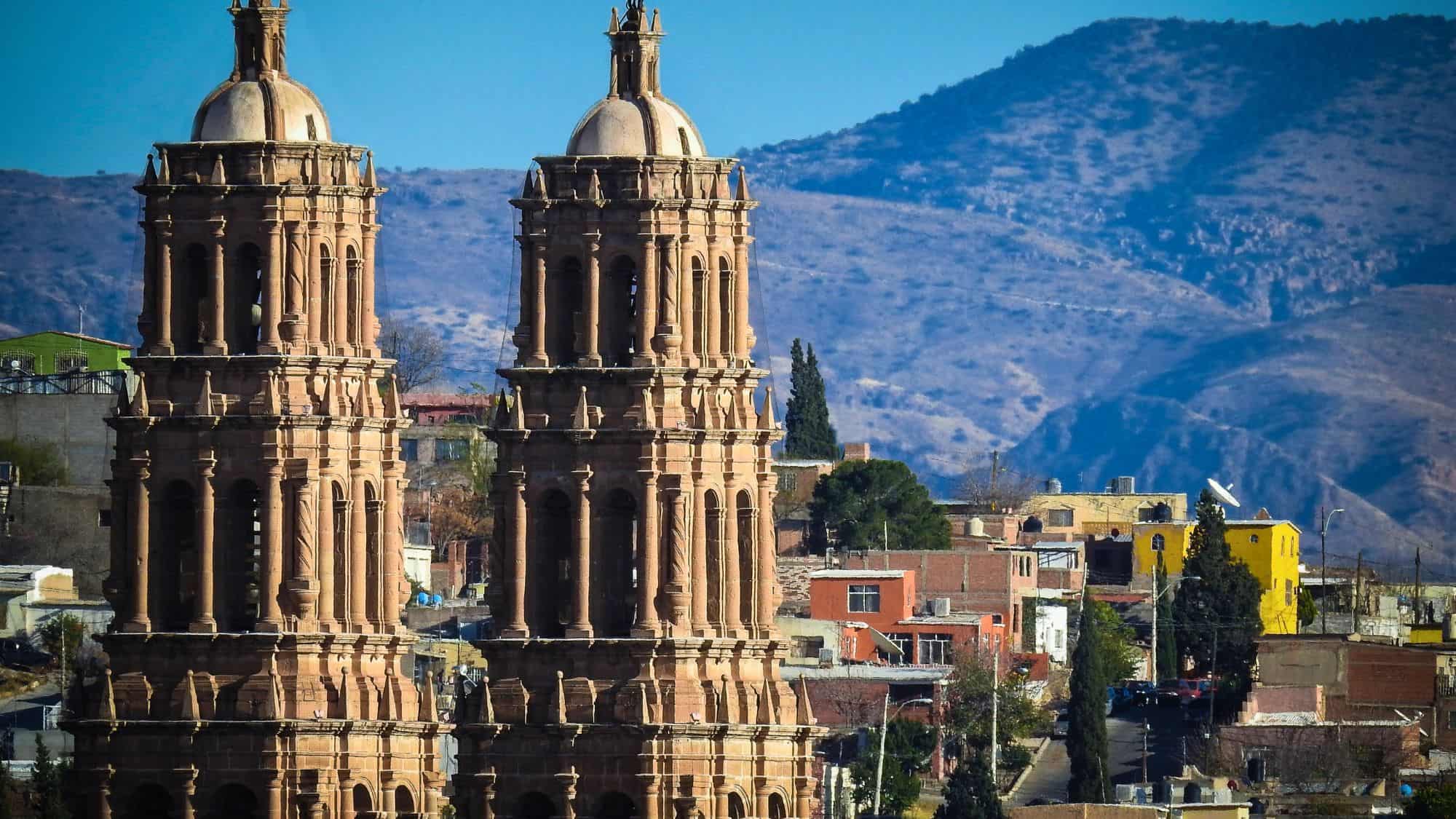 A grand shot of the Cathedral of Chihuahua, with its twin towers standing against a backdrop of rolling hills. The warm tones of the historic stone architecture contrast with the blue sky and the colorful buildings in the foreground.