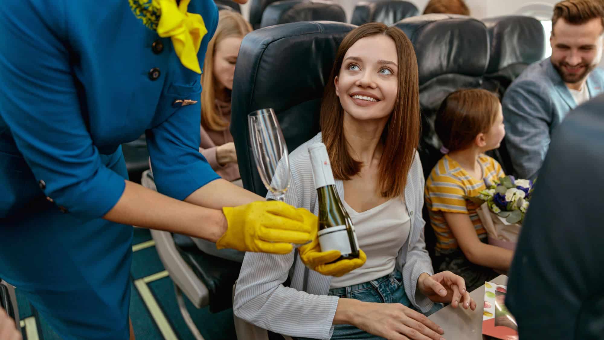A flight attendant in a blue uniform with yellow gloves hands a smiling passenger a bottle of champagne and an empty glass. Other passengers, including a child holding flowers, sit nearby, enjoying the flight.