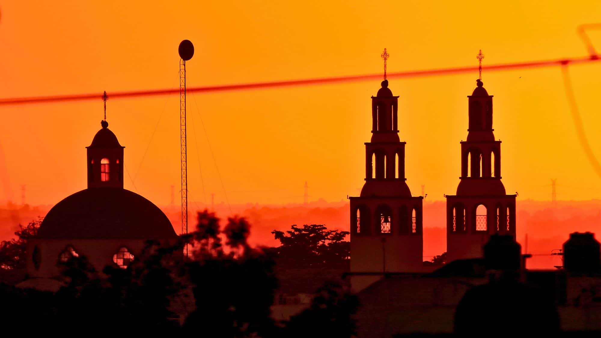 A dramatic silhouette of the Church of Our Lady of San Juan de los Lagos against a fiery orange sunset. The twin bell towers stand tall, creating an iconic and mystical skyline over the city.