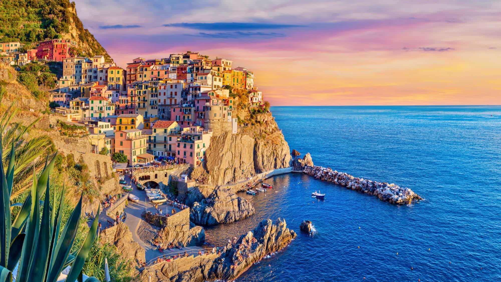 The vibrant coastal village of Manarola, Cinque Terre, Italy, with colorful houses perched on cliffs above the blue Mediterranean Sea, glowing under a pastel sunset sky.